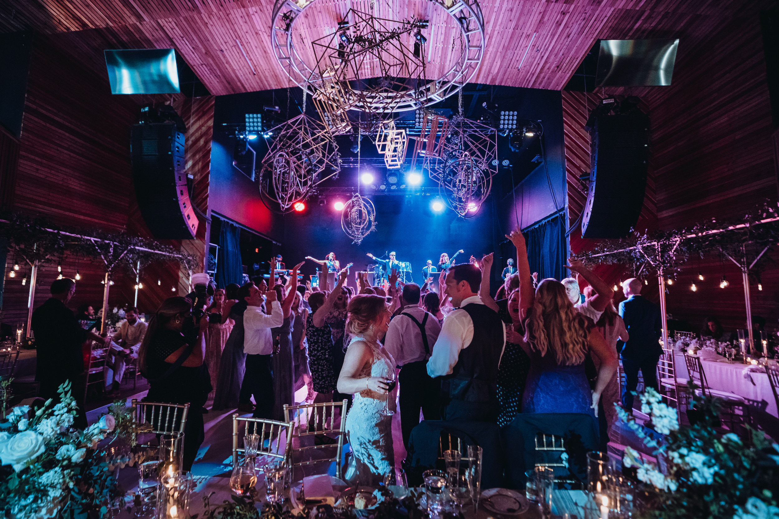 A lively wedding reception with blue, purple and red stage lights and wedding guests dancing to a live band performing on stage at a music hall in Houston, TX.