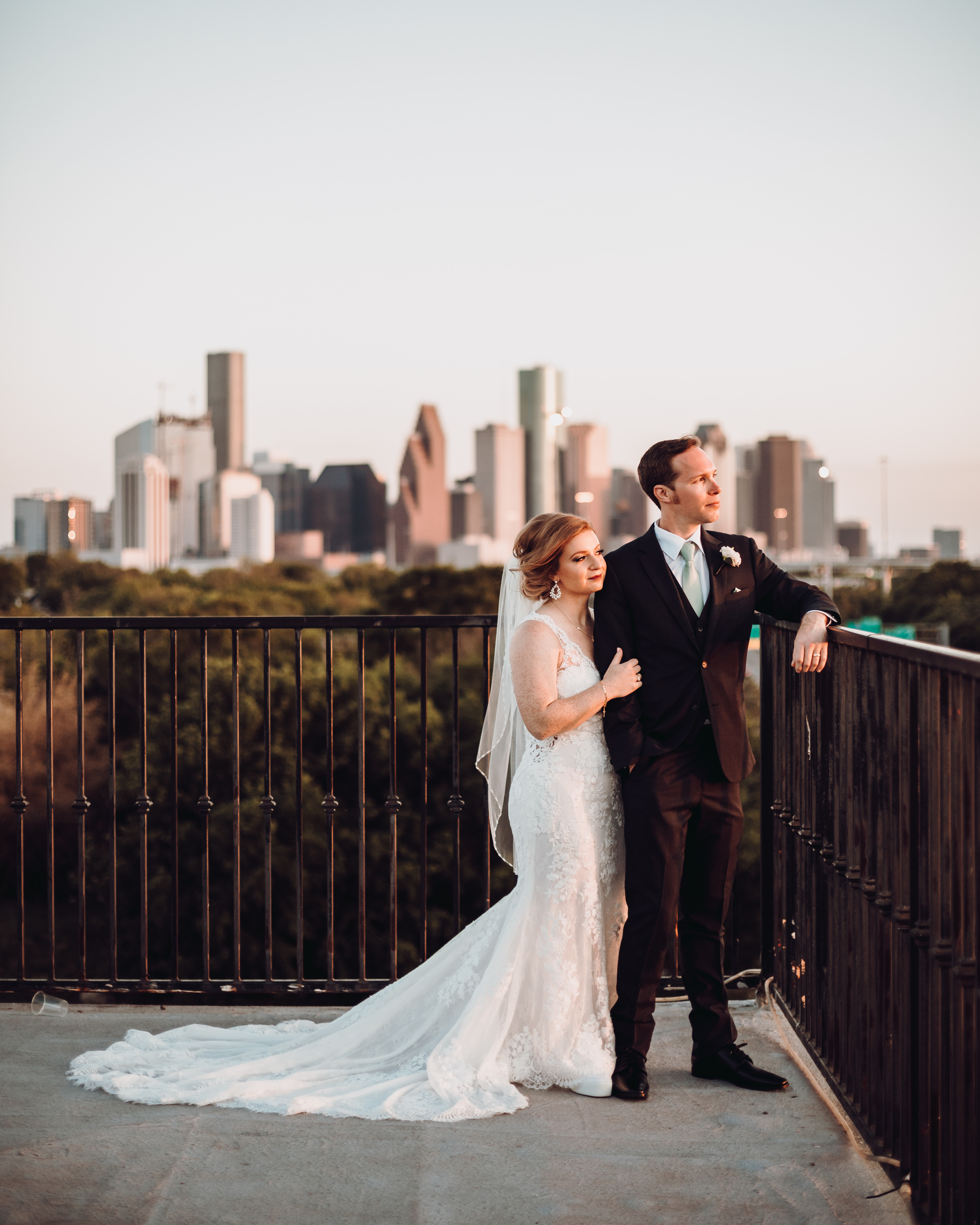 A bride and groom stand outside on a balcony in front of the Houston skyline at their wedding venue.