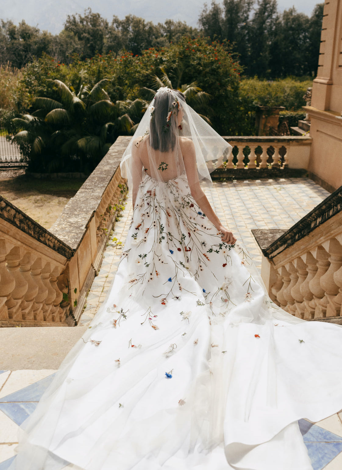 A bride wears a Monique Lhuillier wedding gown that is short and fitted with a flowing train. The strapless wedding dress has colorful embellished flowers covering it.