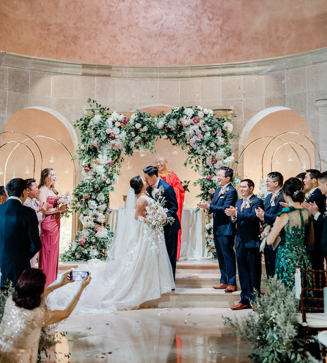The Bell Tower on 34th, a wedding venue in Houston, TX has an onsite chapel. A bride and groom kiss at the altar while the guests stand.