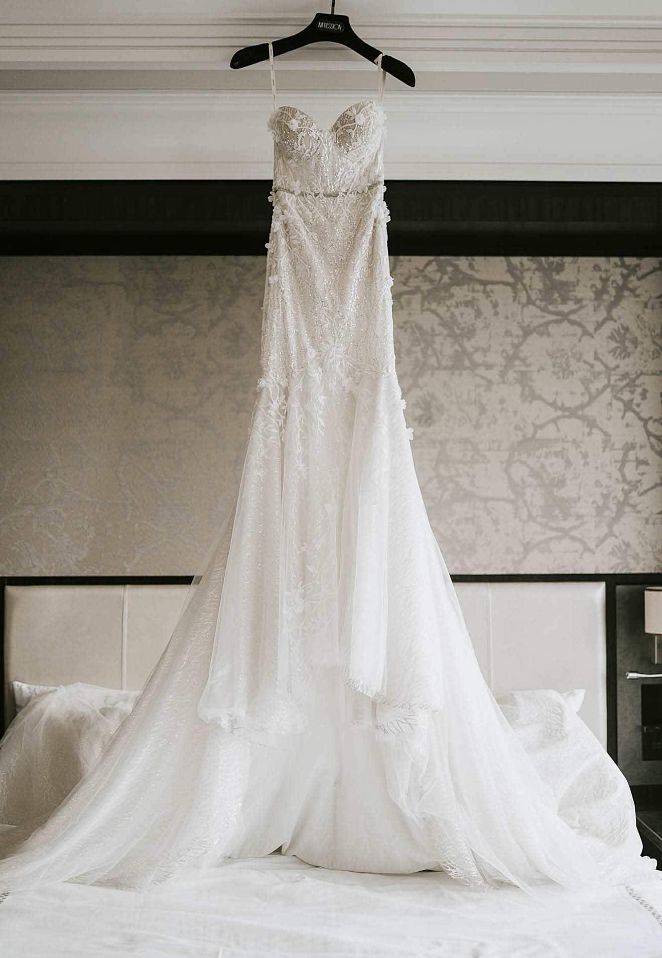 A bride's dress hangs above a hotel bed before the wedding. The dress is a Berta gown and has sparkling flower-embellished details that trail down the skirt. 