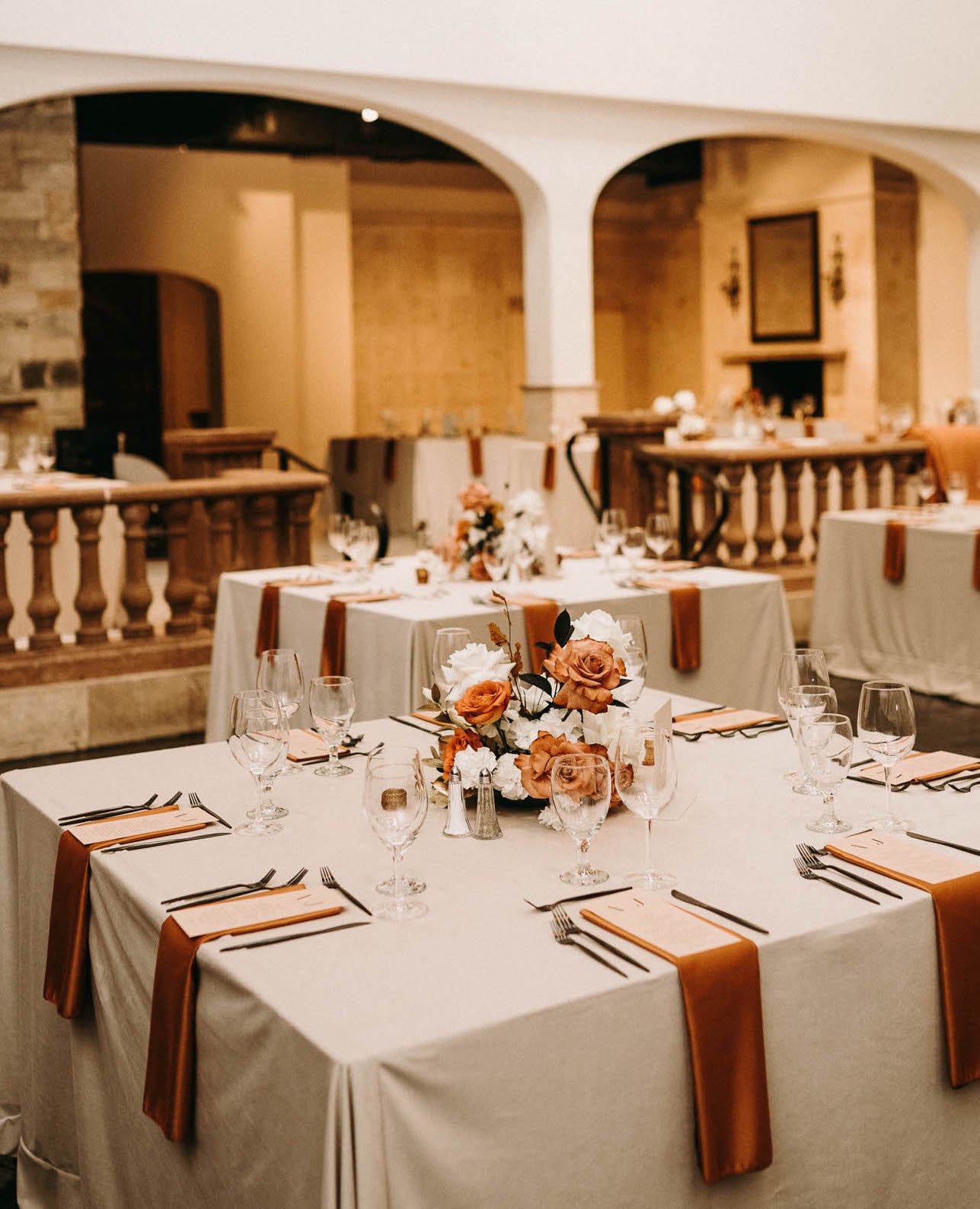 A wedding reception is decorated with burnt orange accents and centerpieces with white and orange flowers. The reception is taking place at The Bell Tower on 34th in Houston, TX.