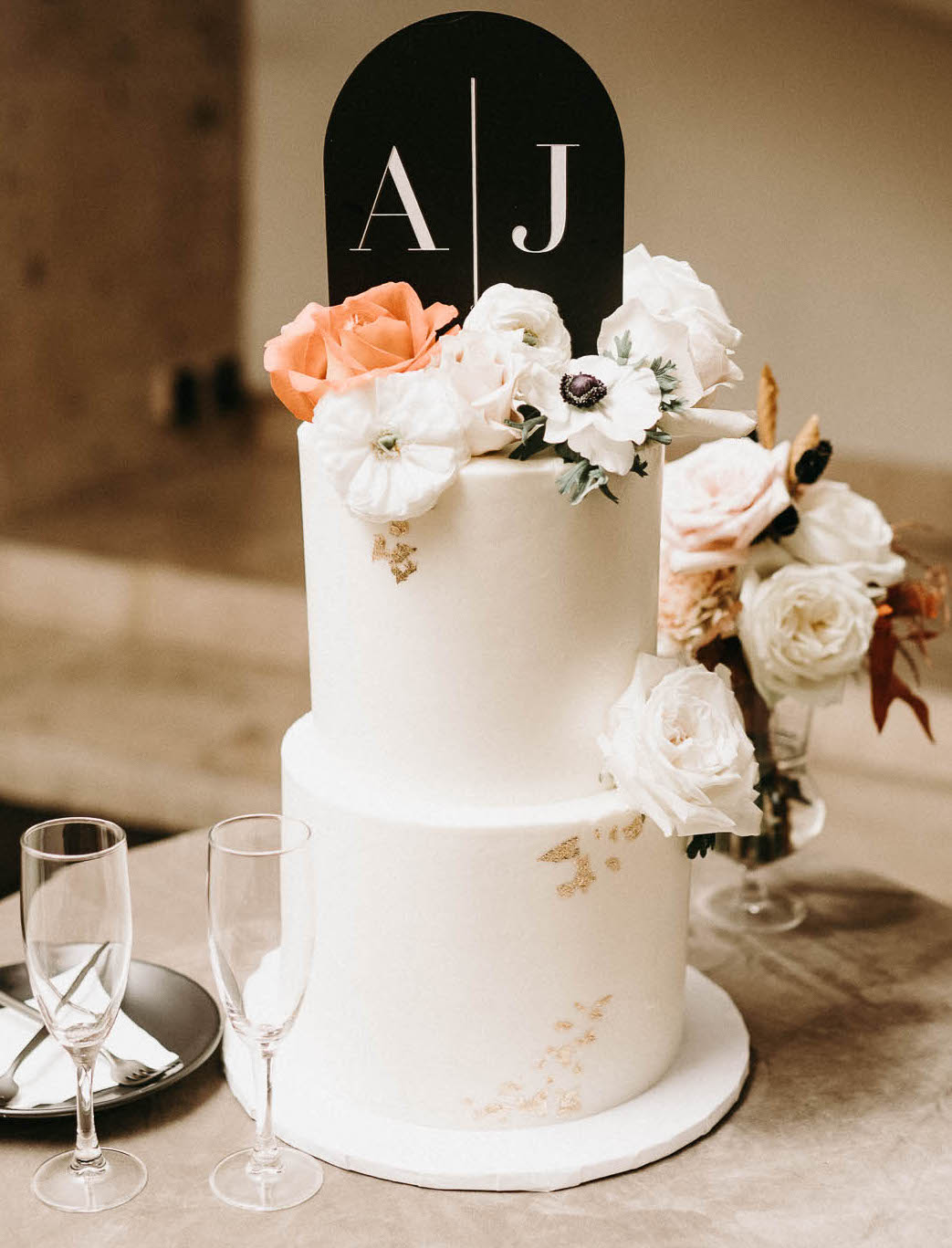 A two-tier white wedding cake with gold flakes and white and peach cake flowers with a black cake topper with the bride and groom's initials.