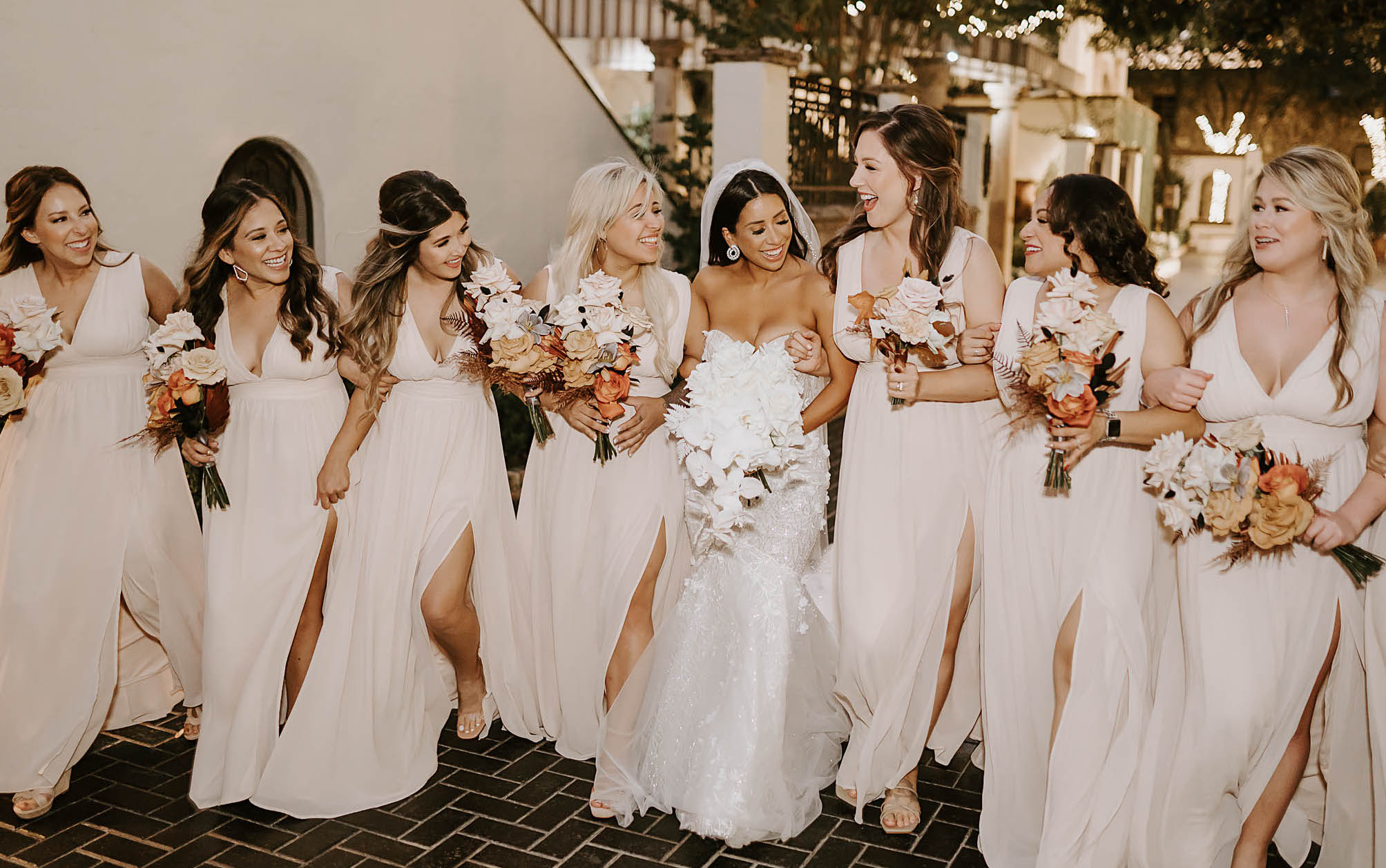 A bride smiles and stands in the middle of all of her bridesmaids before her wedding. All of them are holding bouquets with orchids and burnt-orange flowers.