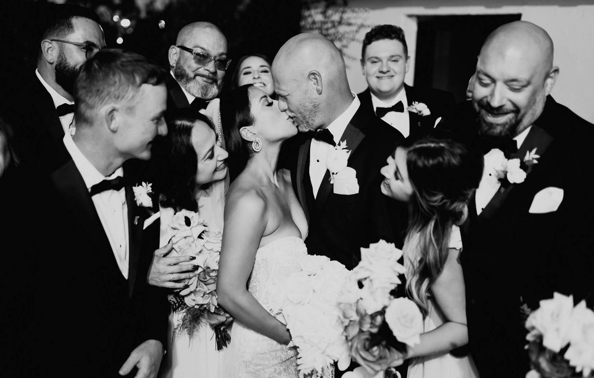 A black and white photo of a bride and groom kissing while their wedding guests circle around them.