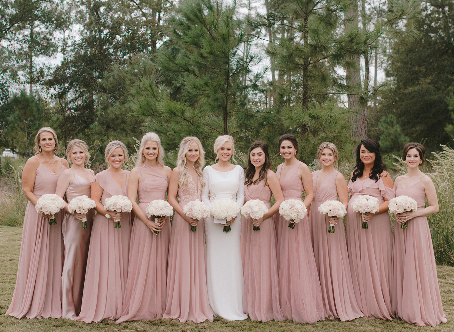 A bride stands with her bridesmaids who are all wearing mauve pink dresses before the bride's romantic ballroom wedding at The Post Oak Hotel.