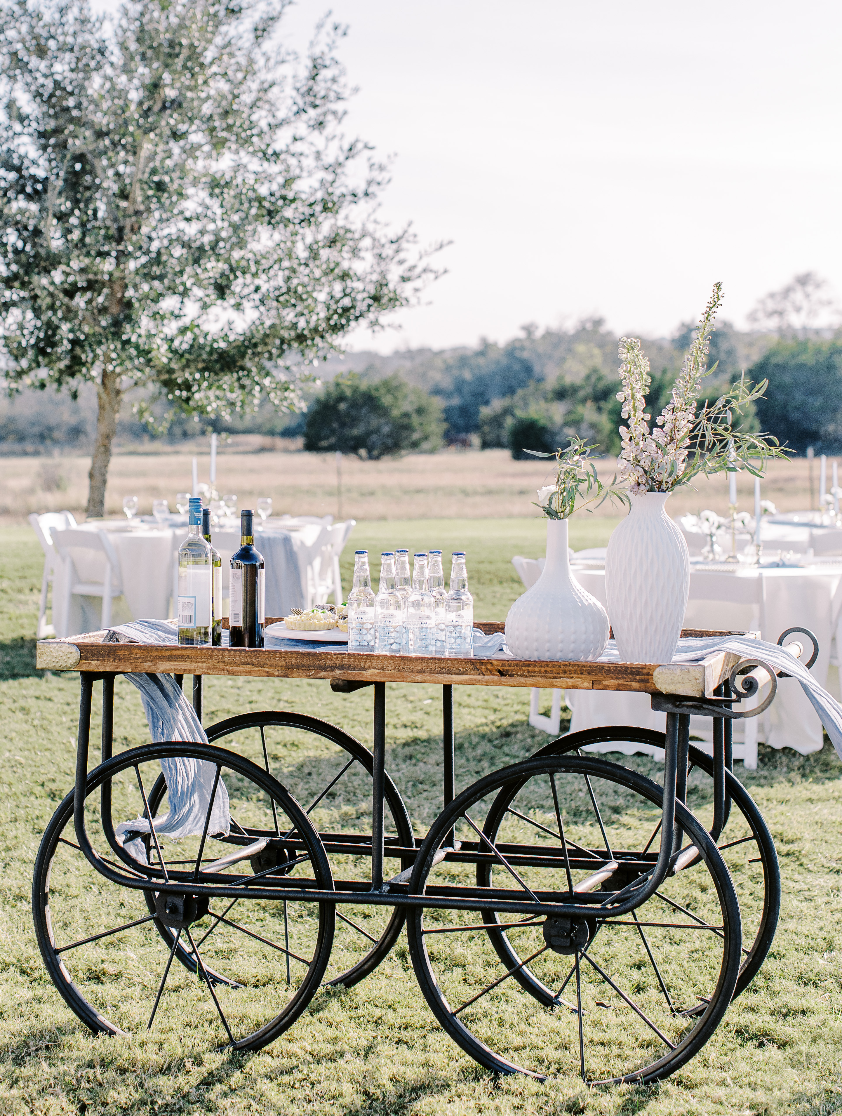 A vintage bar cart with large steel wheels is set up outside for a sunlit hill country wedding editorial in Dripping Springs, TX at Cricket Hill Ranch.