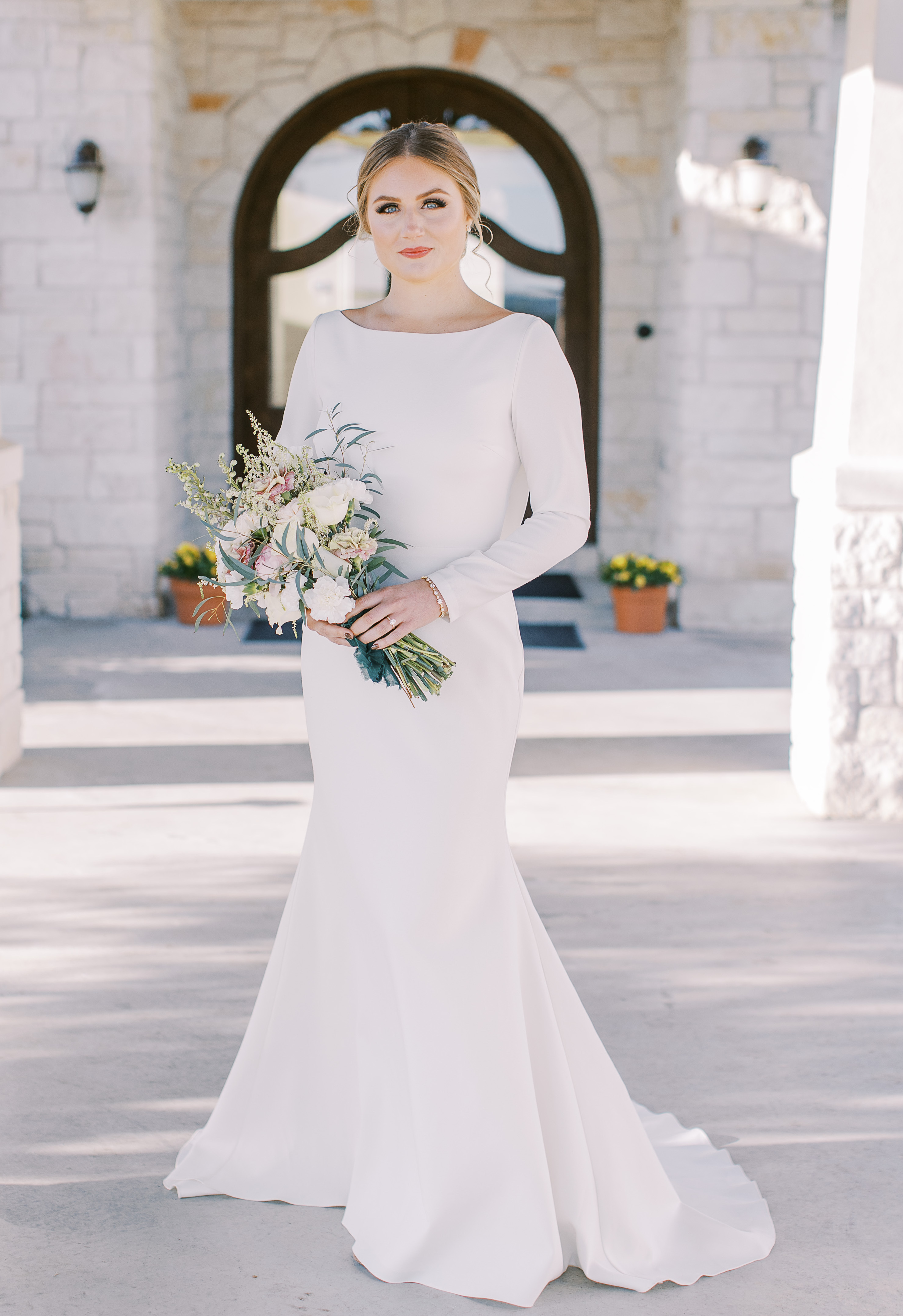 A bride wears a white long sleeve wedding dress and holds a bridal bouquet during a sunlit hill country editorial in Dripping Springs, TX.