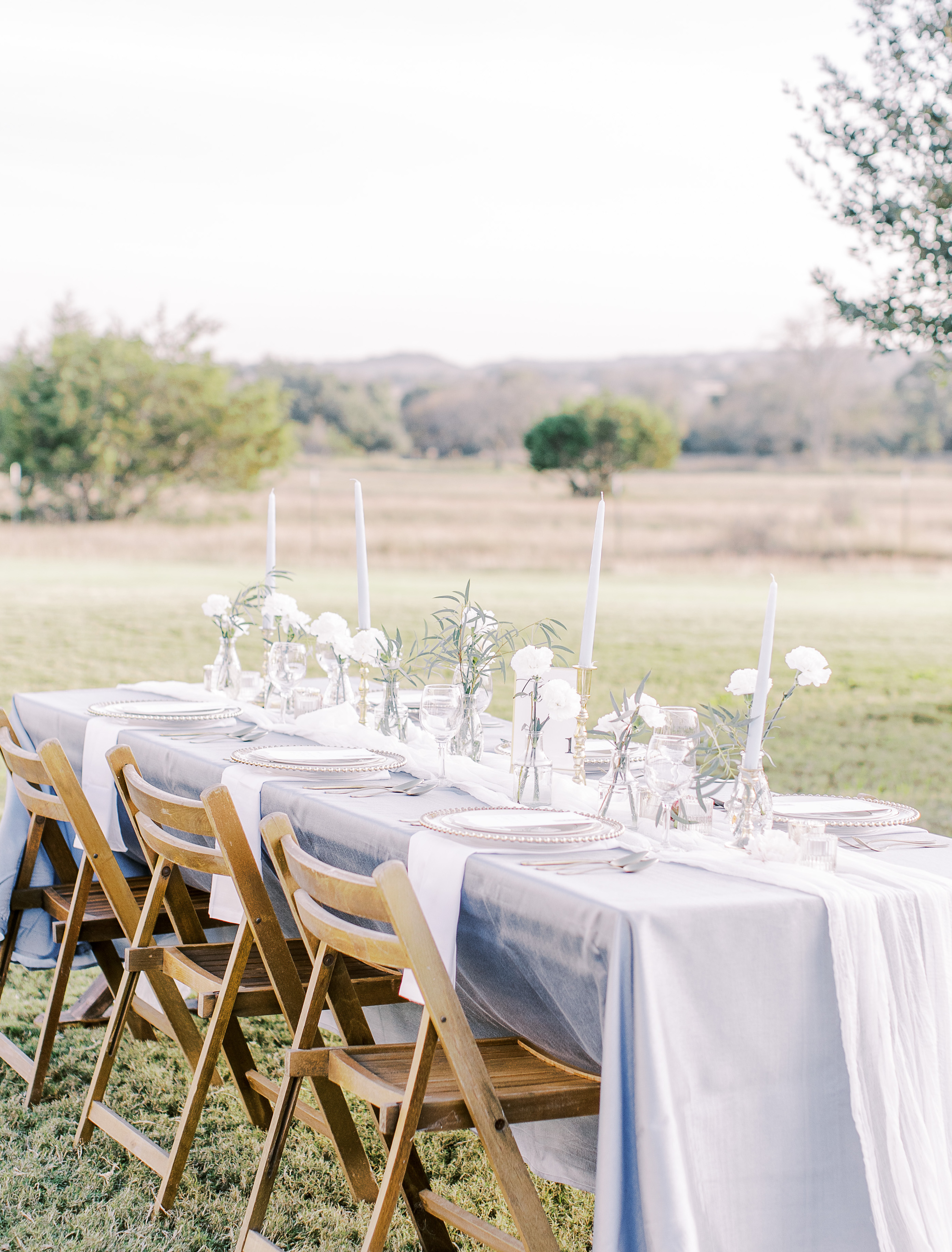 An intimate wedding reception is alfresco with a family-style table decorated with light blue taper candles and white flowers in bud vases.