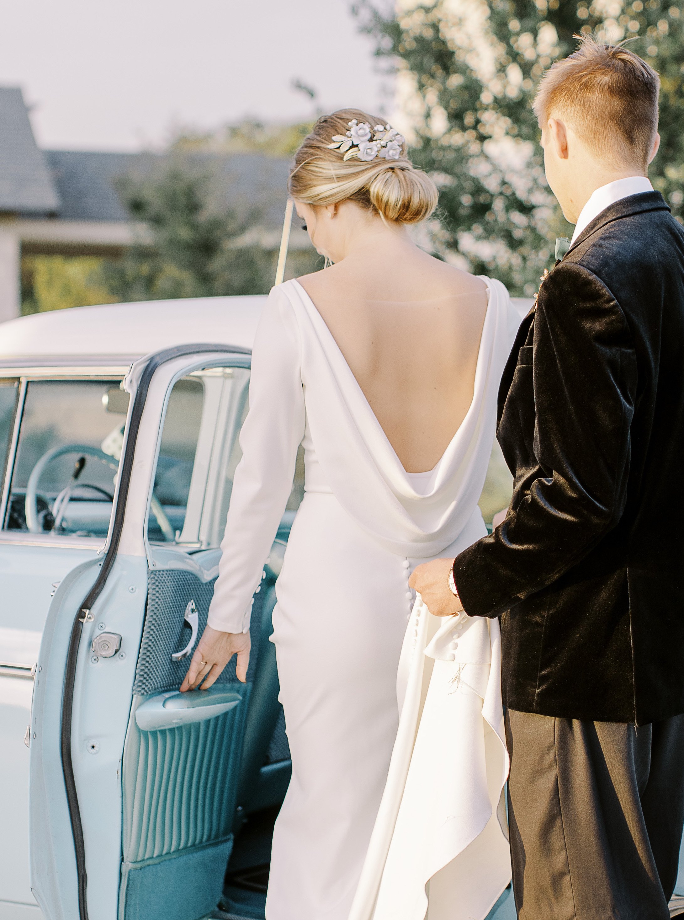 A groom picks up with train of a brides gown as she gets in the backseat of a vintage light blue car.