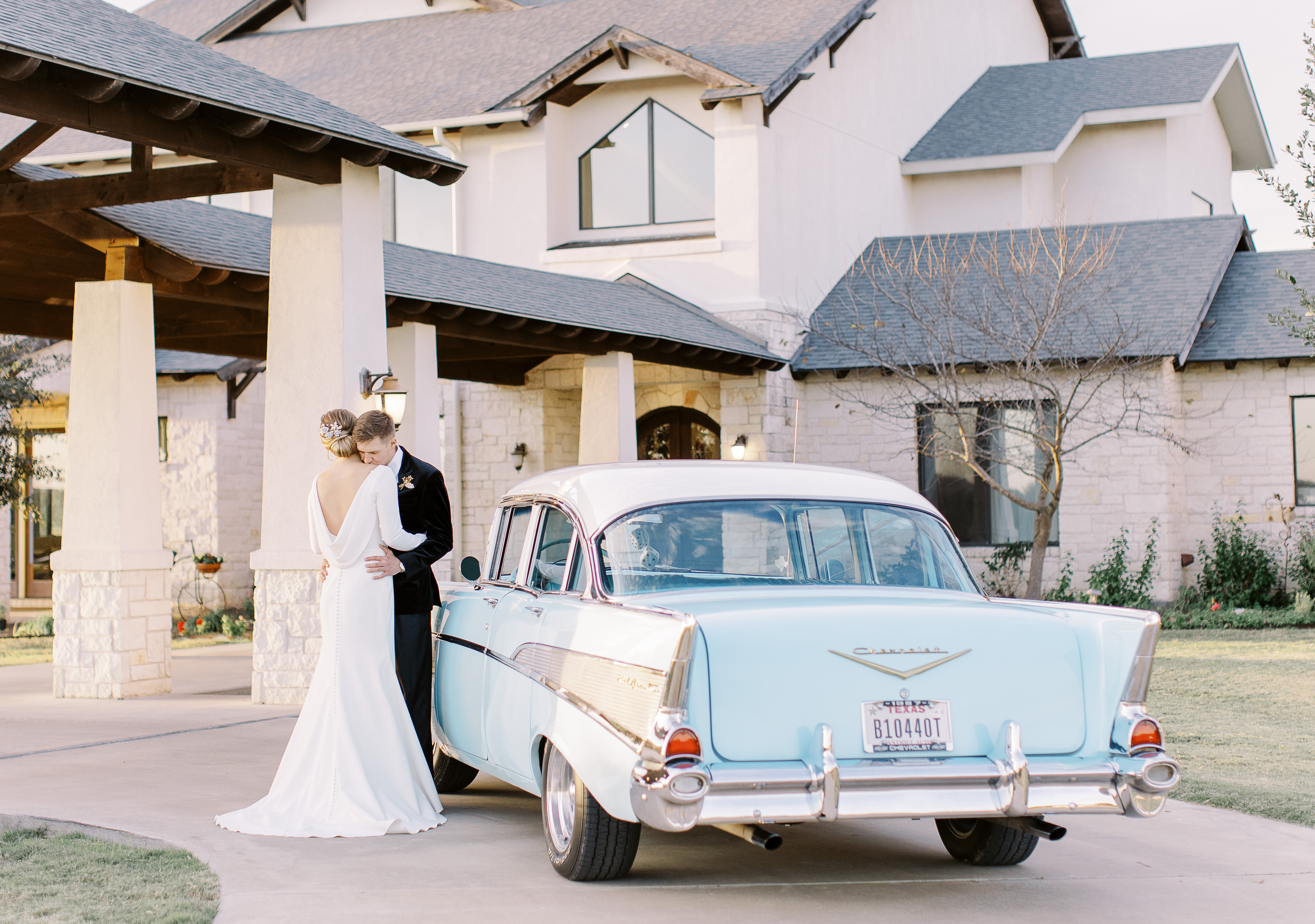 A bride and groom hug in front of the Texas Hill Country venue Cricket Hill Ranch. A vintage baby blue car is parked beside them.