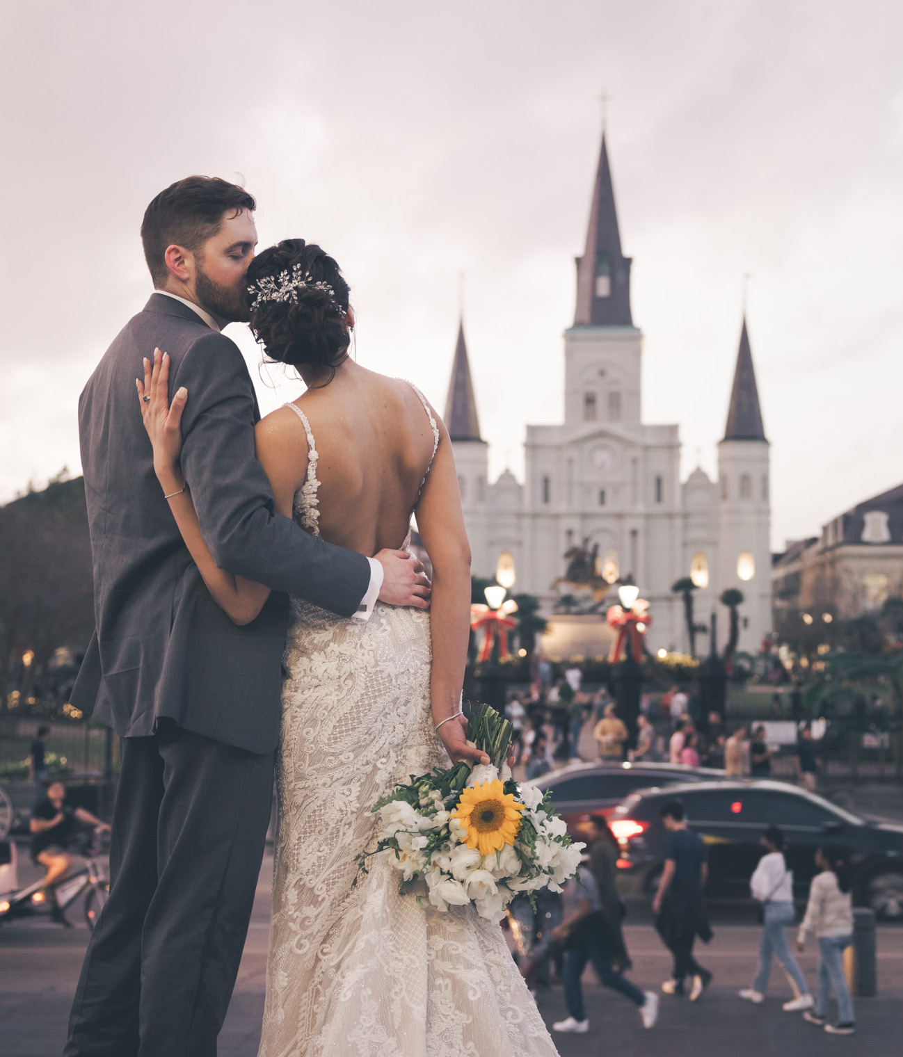 A groom kisses his bride on the head as they look at the busy streets of the French Quarter in New Orleans during their wedding day.