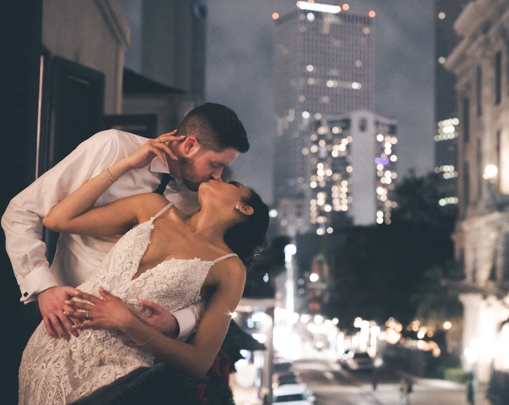 A bride and groom kiss on a balcony at night with the NOLA city lights in the background.
