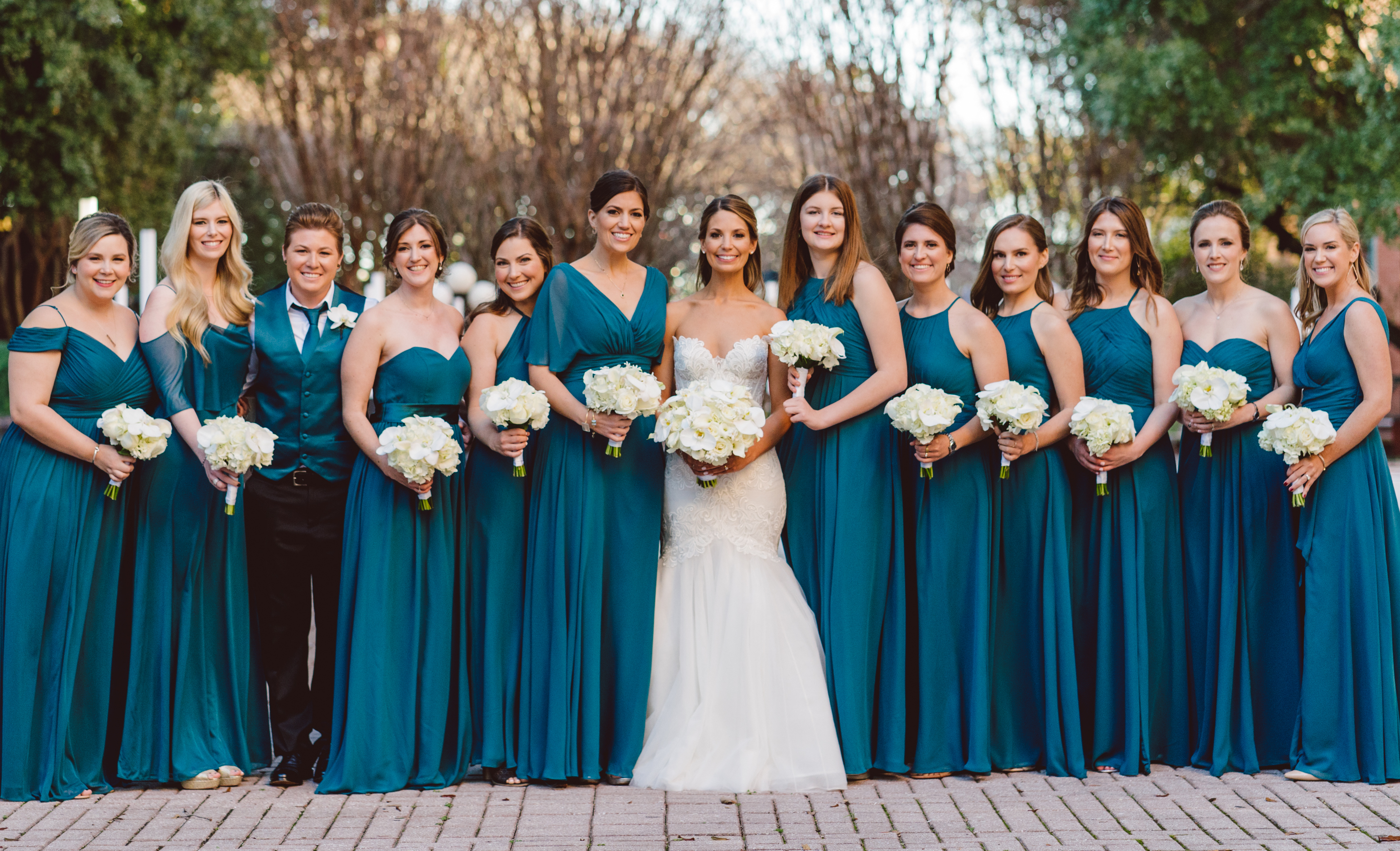 A bride poses with her bridesmaids outside. They are all wearing deep teal dresses for an opulent winter wedding in Houston.