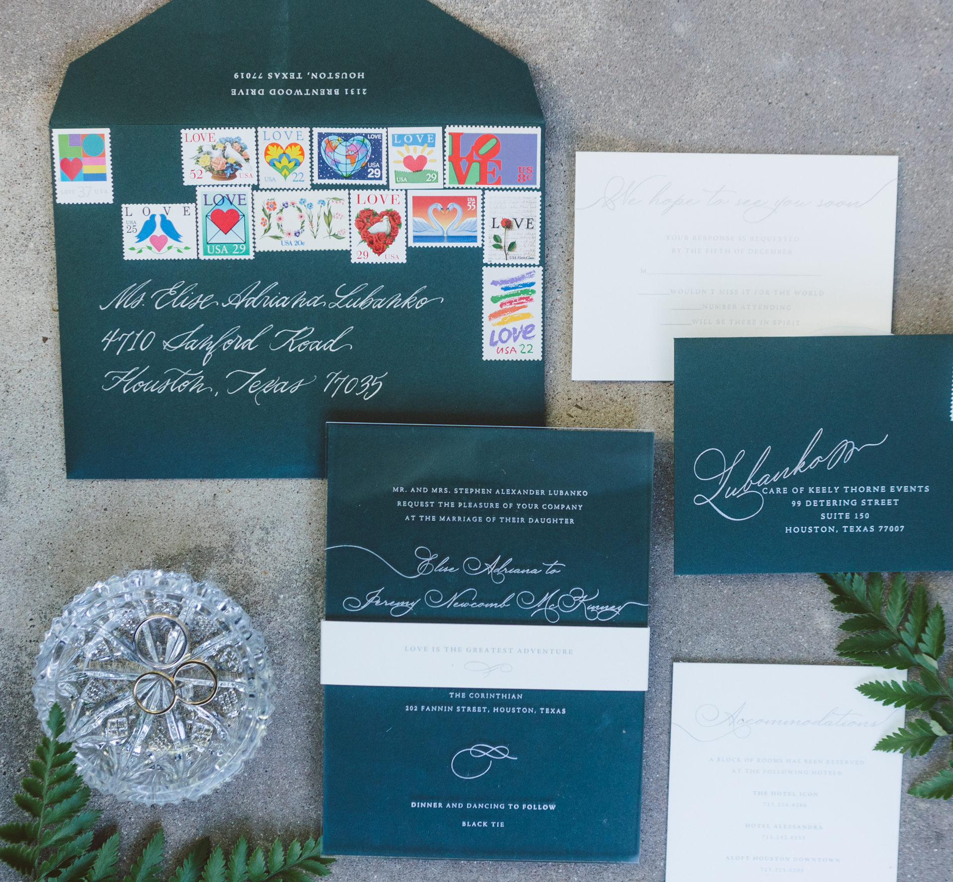 A flatlay of dark teal wedding invitations with cursive white writing and green leaves bordering them.