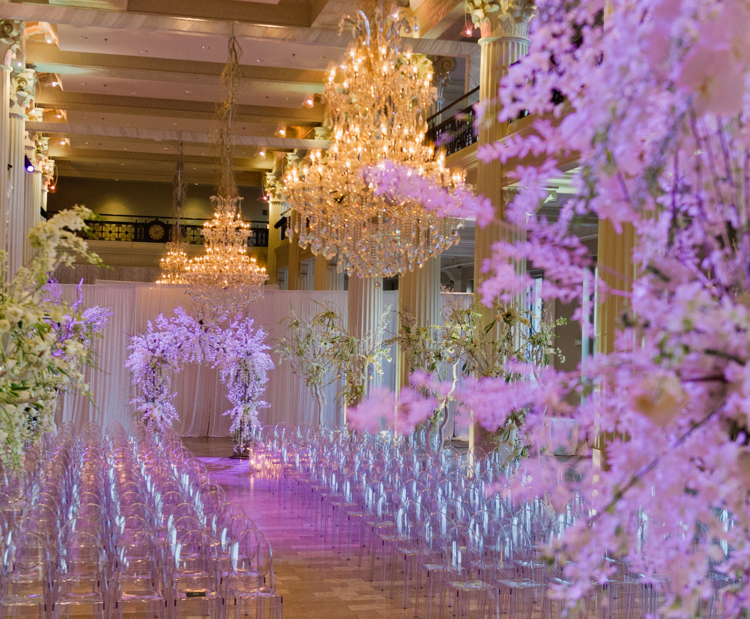 A ceremony space is decorated with crystal chandeliers and white flower installations for a winter wedding in Houston. The room is illuminated with a magenta light.