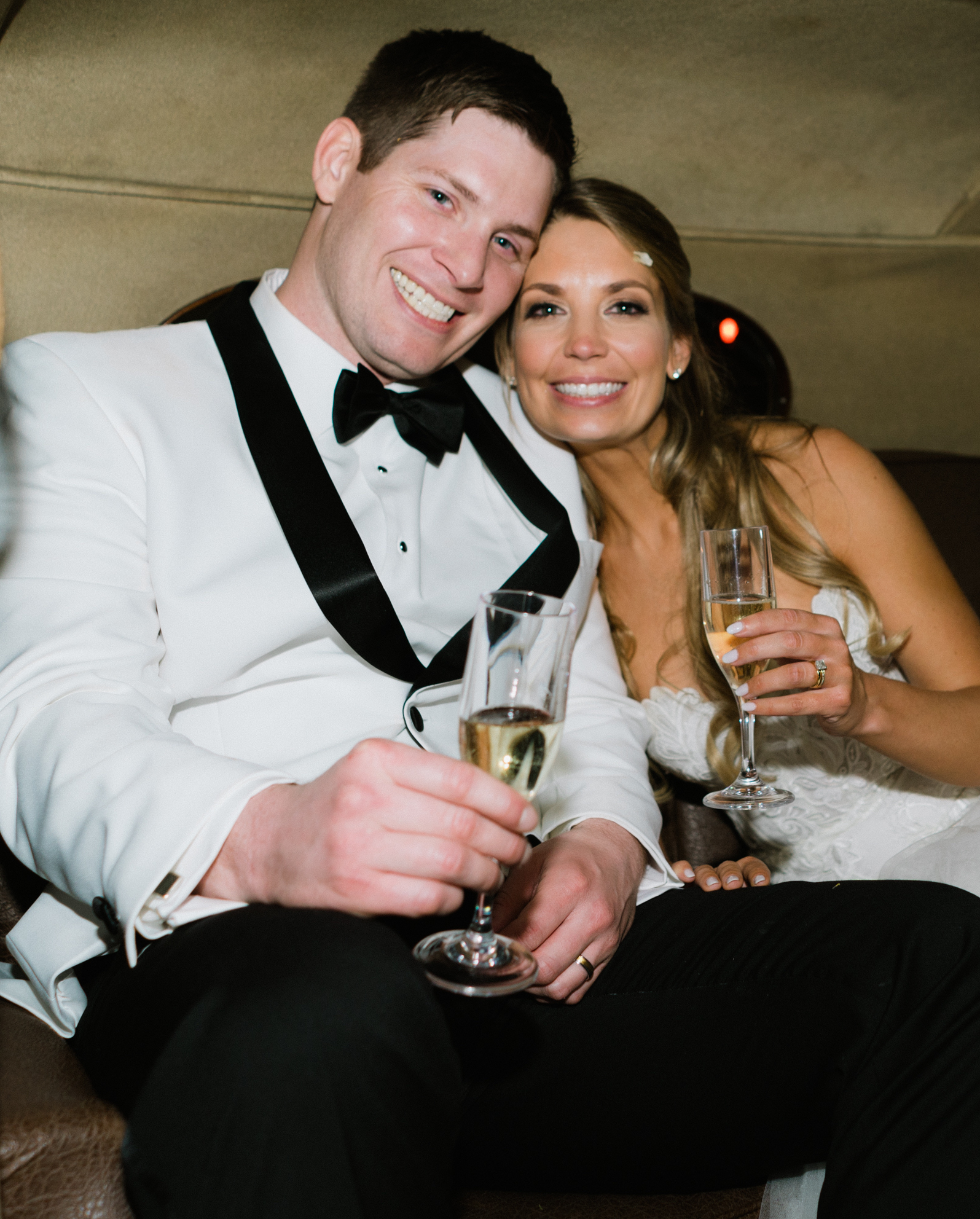 A bride and groom sit in the backseat of a vintage car with glasses of champagne in hand.