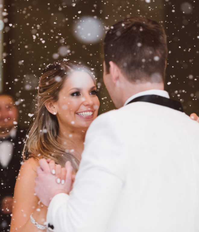 Bride smiles at a groom while dancing while fake snows falls from the ceiling at their wedding reception at the corinthian houston. 