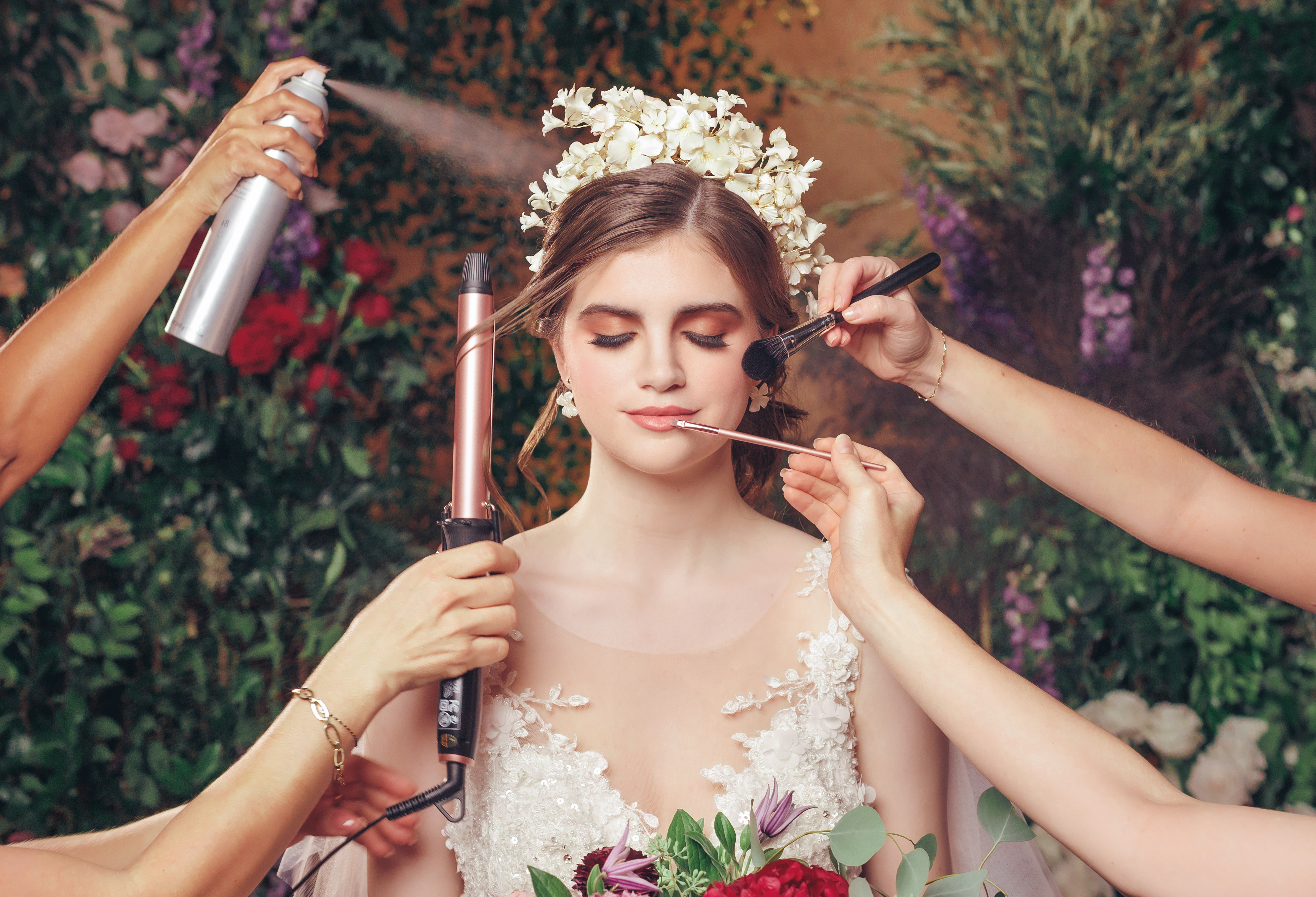 Four hands with hairspray, a curling iron, lipstick applicator and blush brush apply makeup to a bride with closed eyes at a wedding styled shoot at The Bell Tower on 34th in Houston, Texas. 