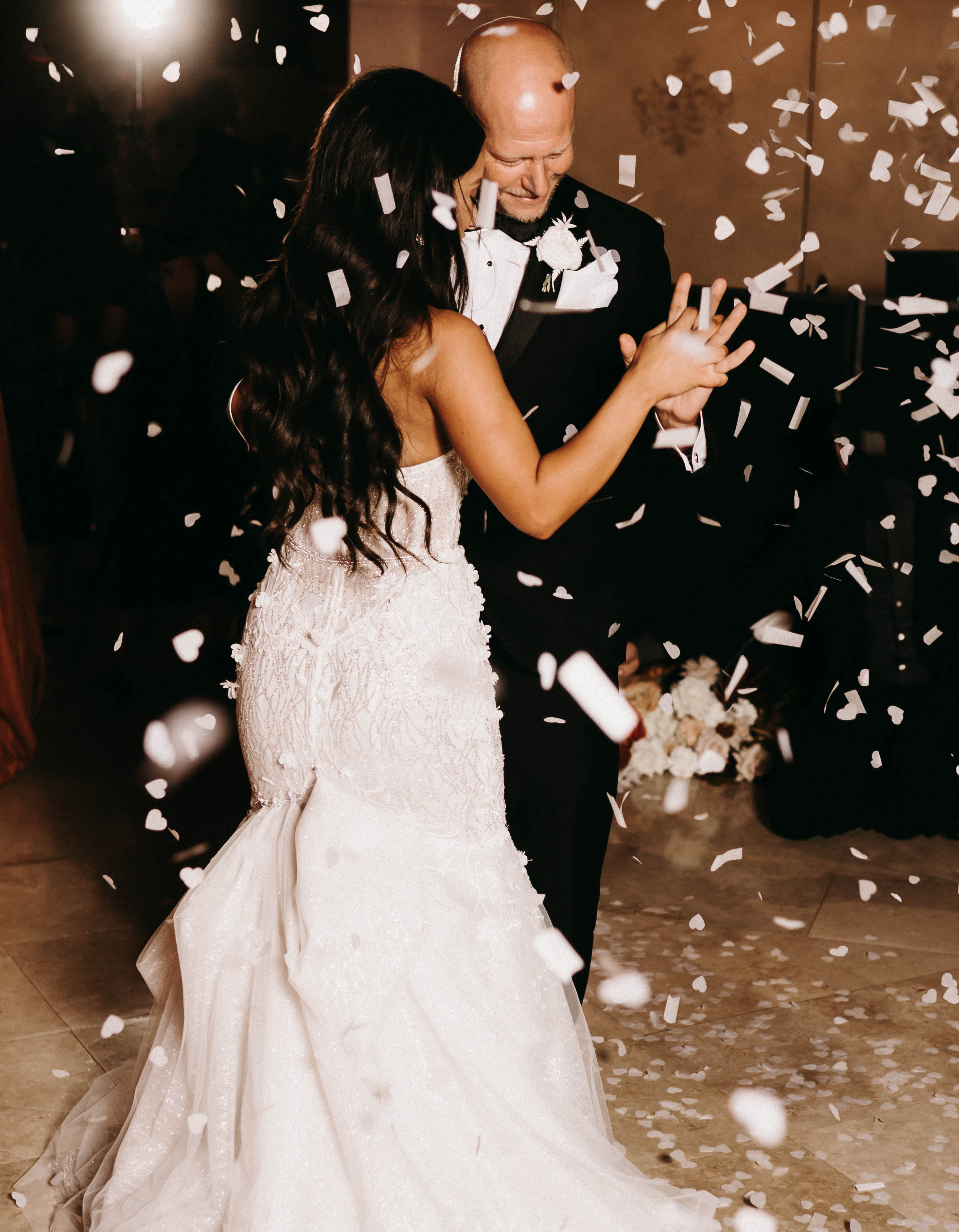 A bride and groom dance at their earth-toned wedding with white confetti falling around them.