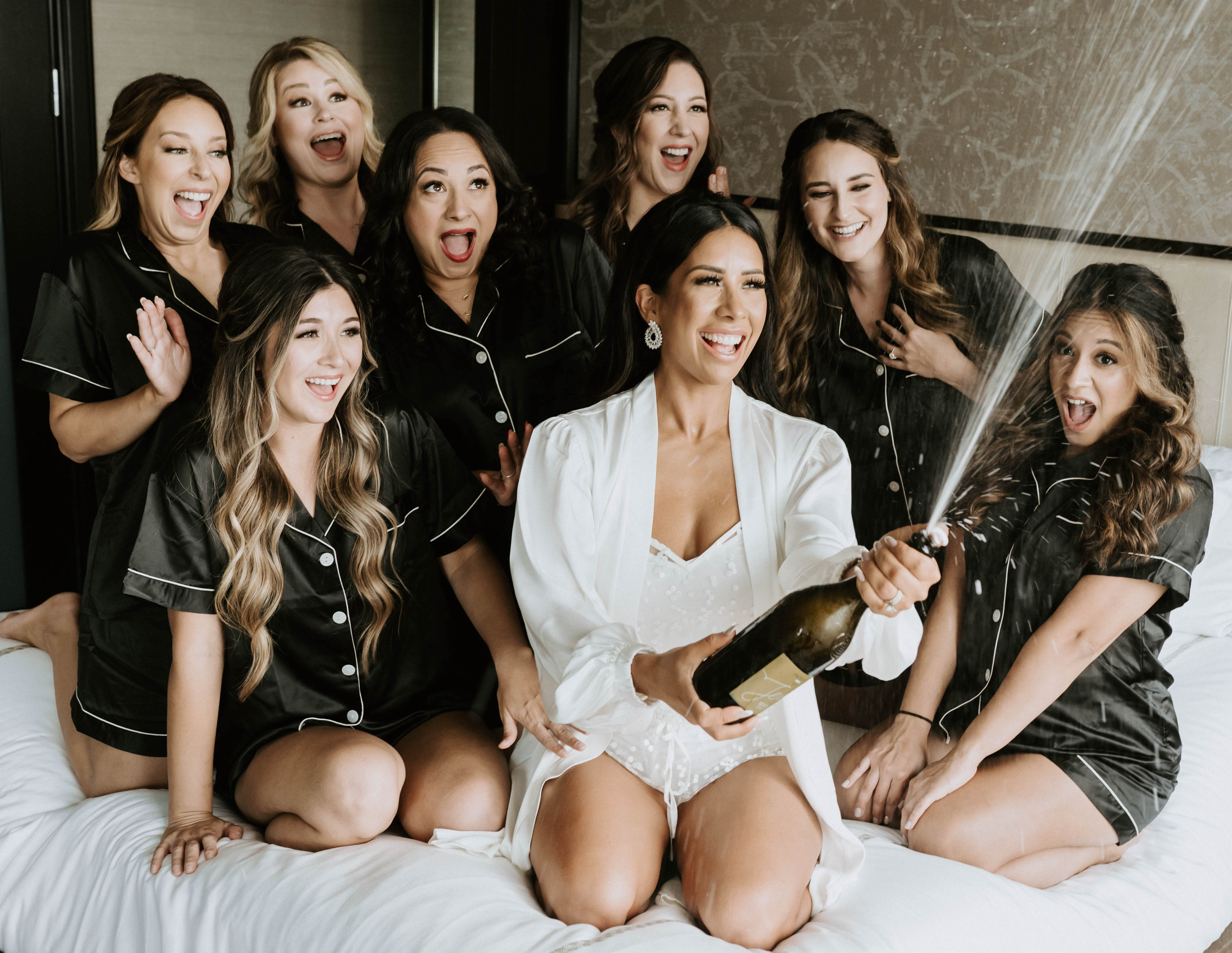 A bride smiles and sprays champagne out of a champagne bottle while sitting on a bed with all of her bridesmaids before her earth-toned wedding at The Bell Tower on 34th in Houston, TX.