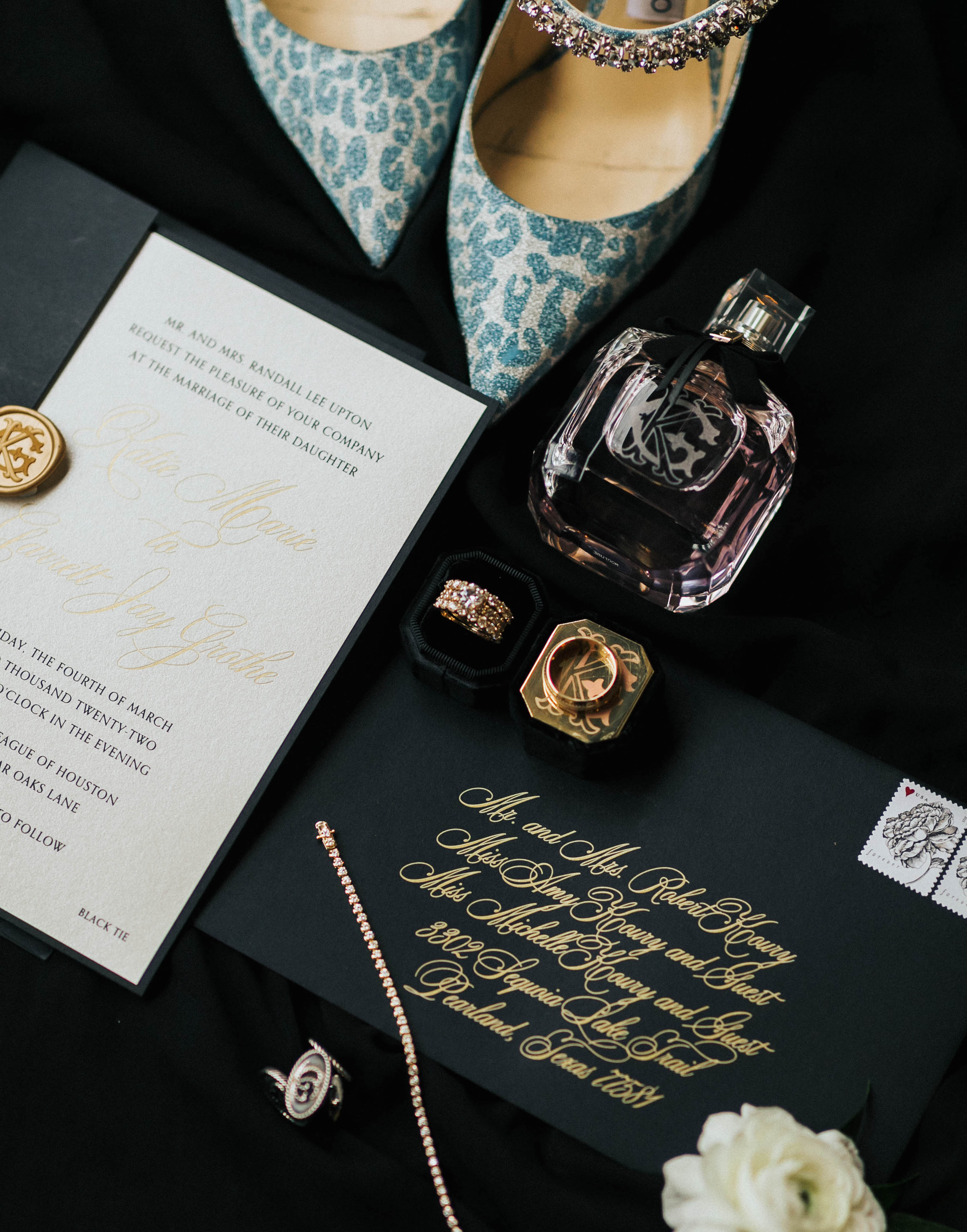 A black wedding invitation suite with a perfume bottle, shoes and wedding bands surrounding the luxury invitations created by Bering's in Houston.