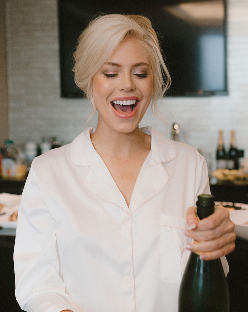 Blonde bride wearing a white button down smiles while holding an open bottle of champagne in a hotel suite at The Post Oak Hotel in Uptown Houston. 