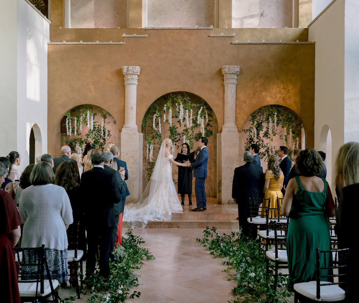 The Bell Tower on 34th, a wedding venue in Houston, TX has an onsite chapel. A bride and groom stand at the altar while the guests stand.