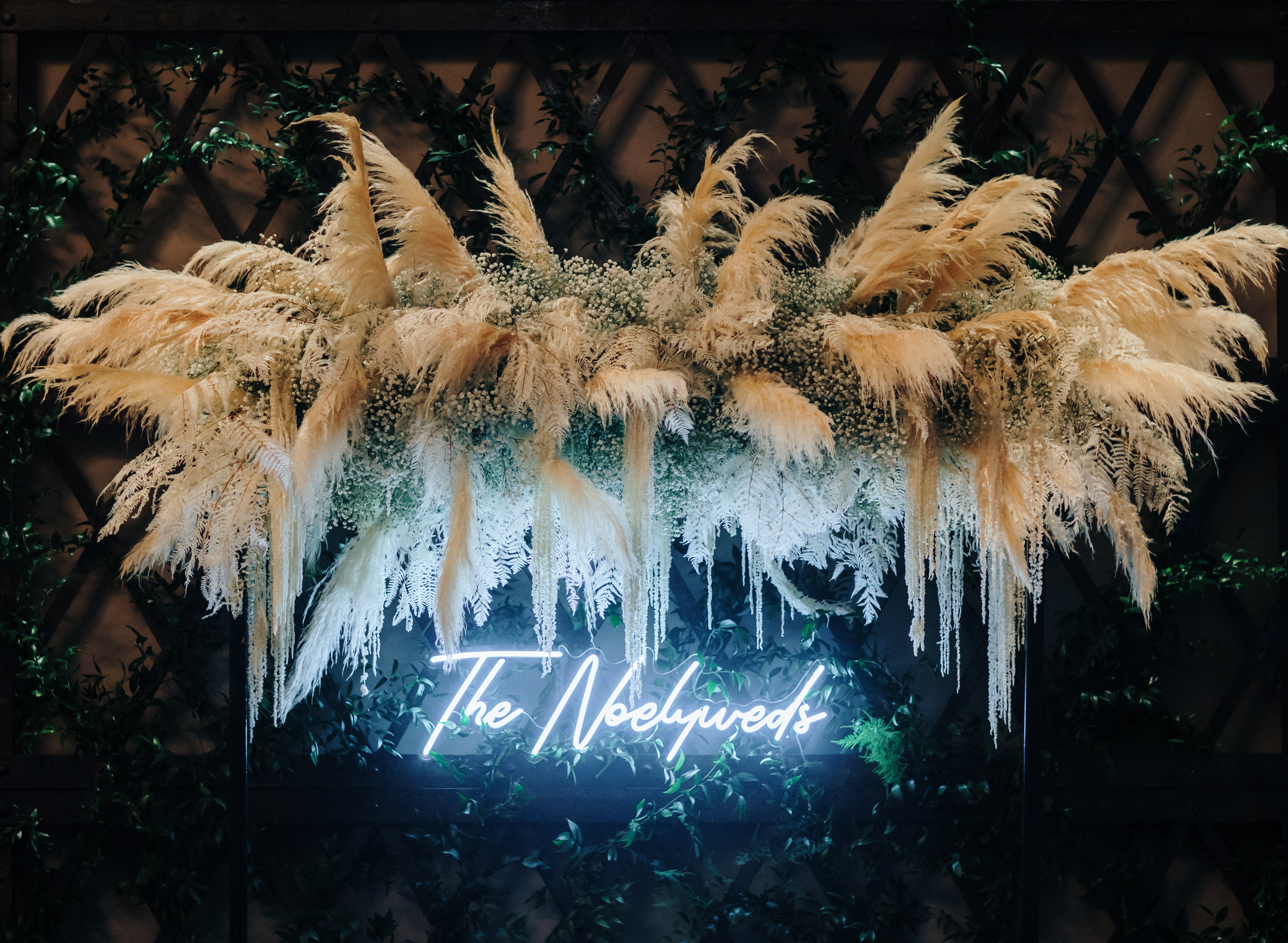A neon sign that says "The Noelyweds" is set up under a floral installation full of baby's breath and pampas grass.