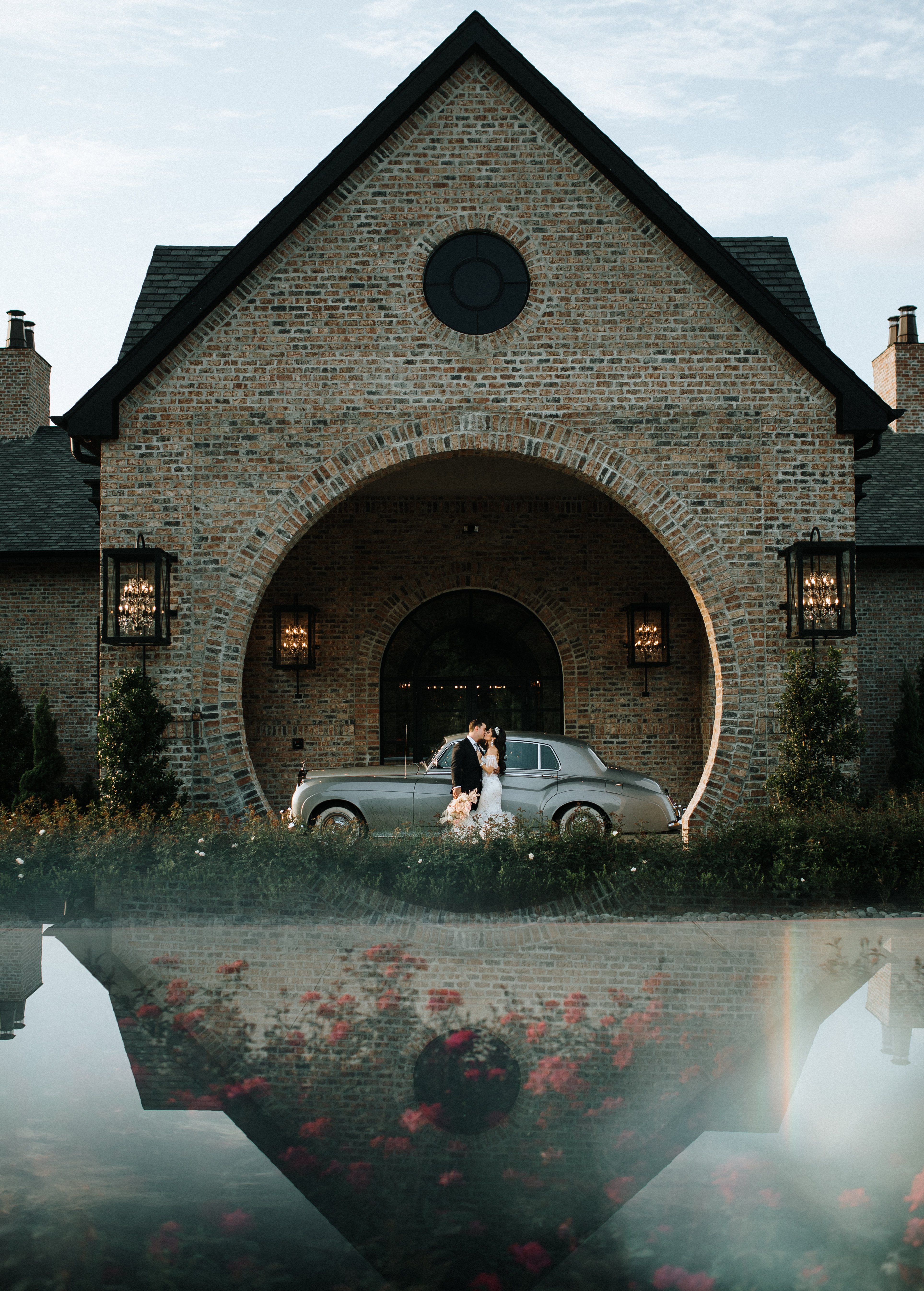 A bride and groom stand in front of silver car and their wedding venue Iron Manor in Montgomery, TX.
