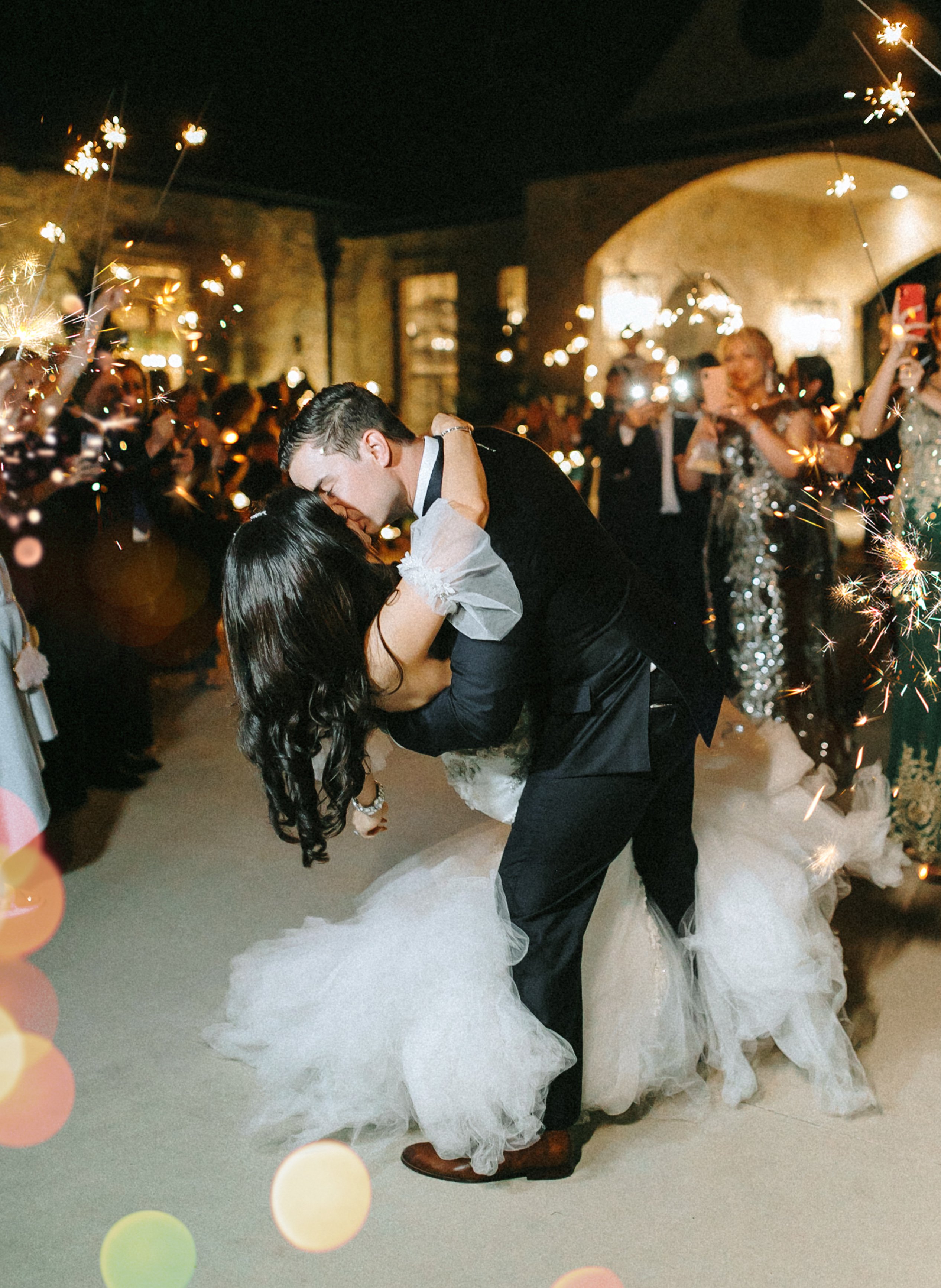 A groom dips his bride in the middle of their guests who are holding out sparklers for their wedding send-off.