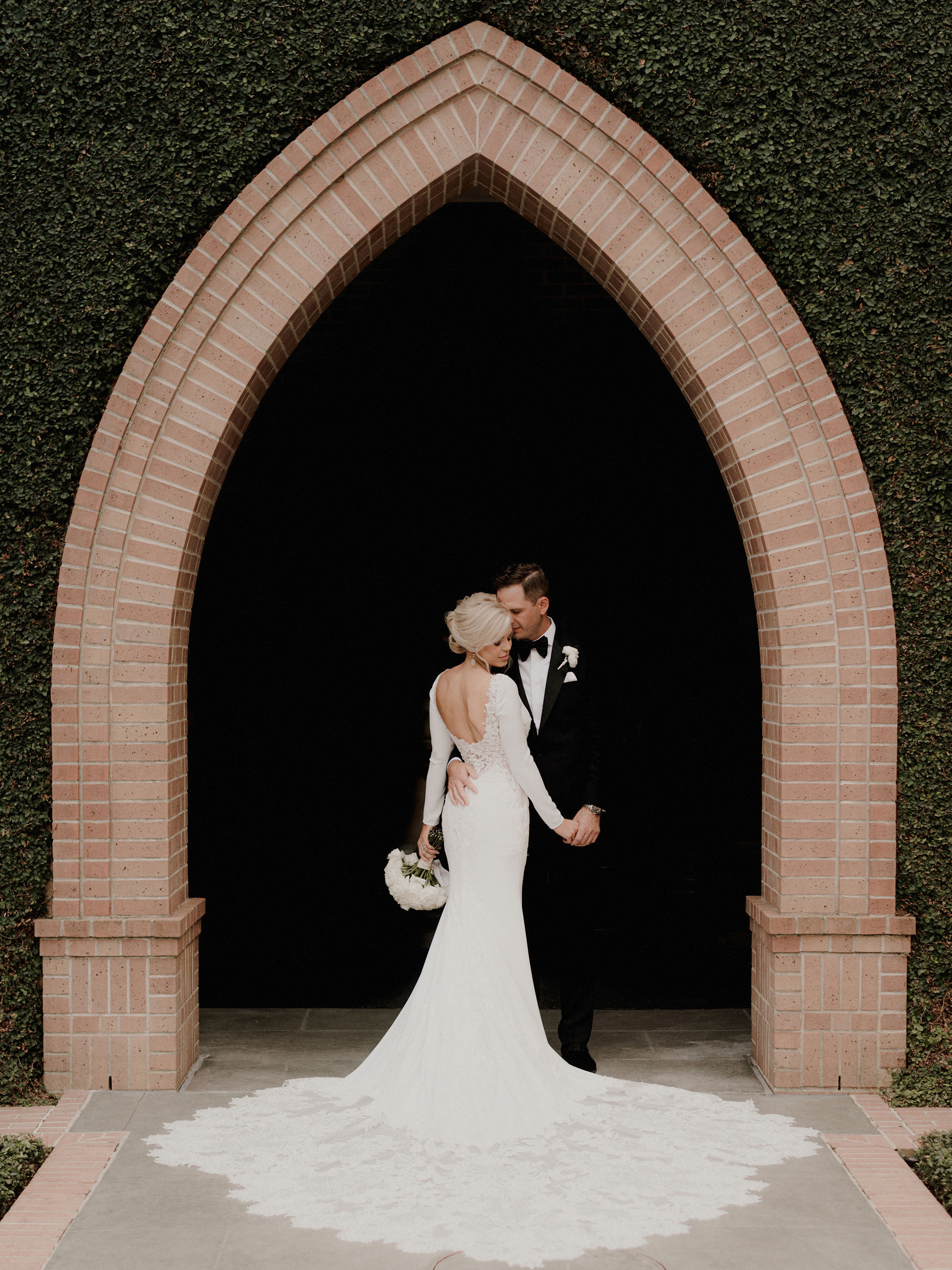 A groom holds a bride underneath a brick arch surrounded by live greenery on their wedding day at St. Marks in Houston, Texas.