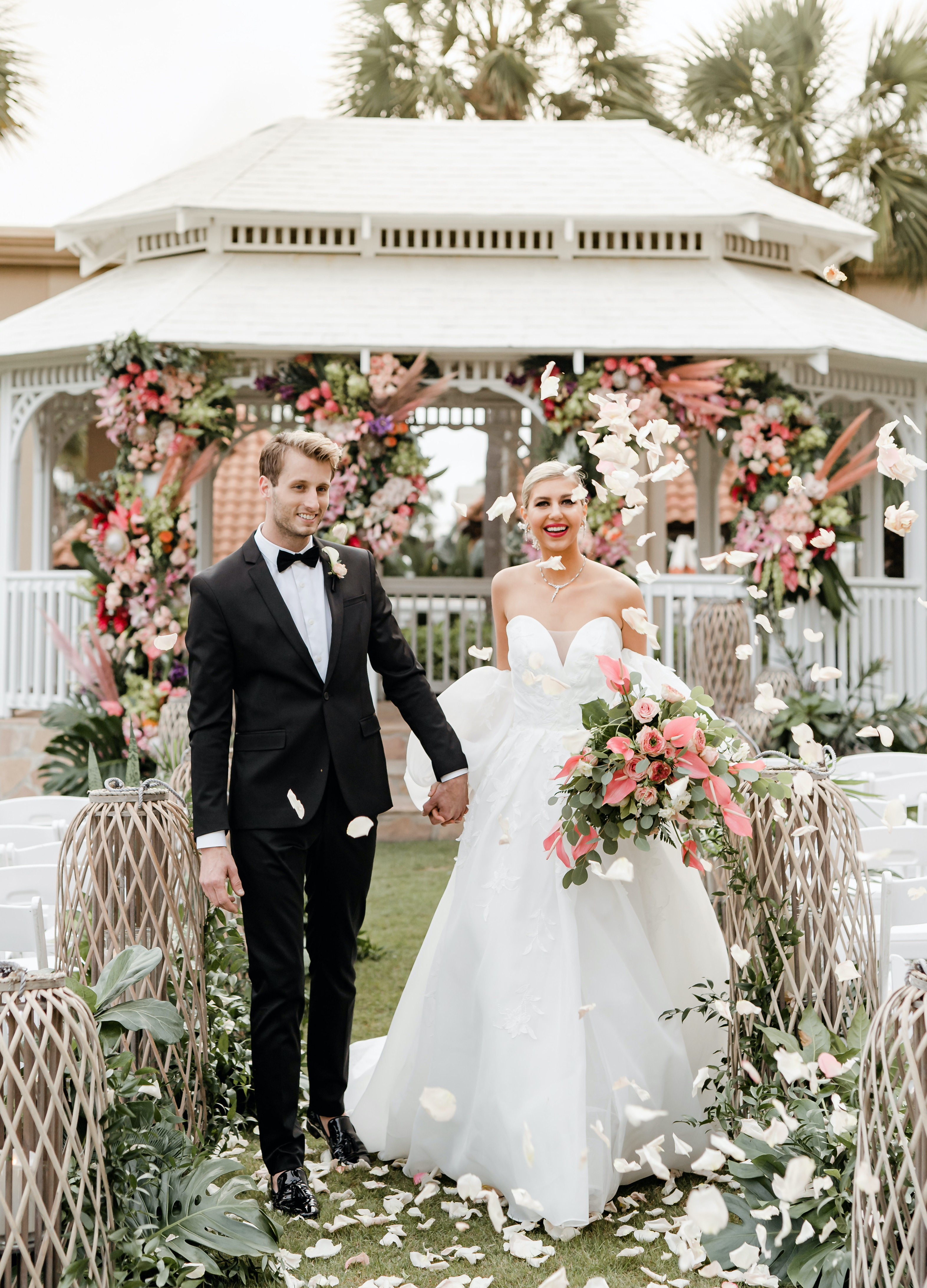 A bride and groom walk down the aisle while rose petals are being thrown in the air around them during a tropical luxe beachfront wedding editorial.