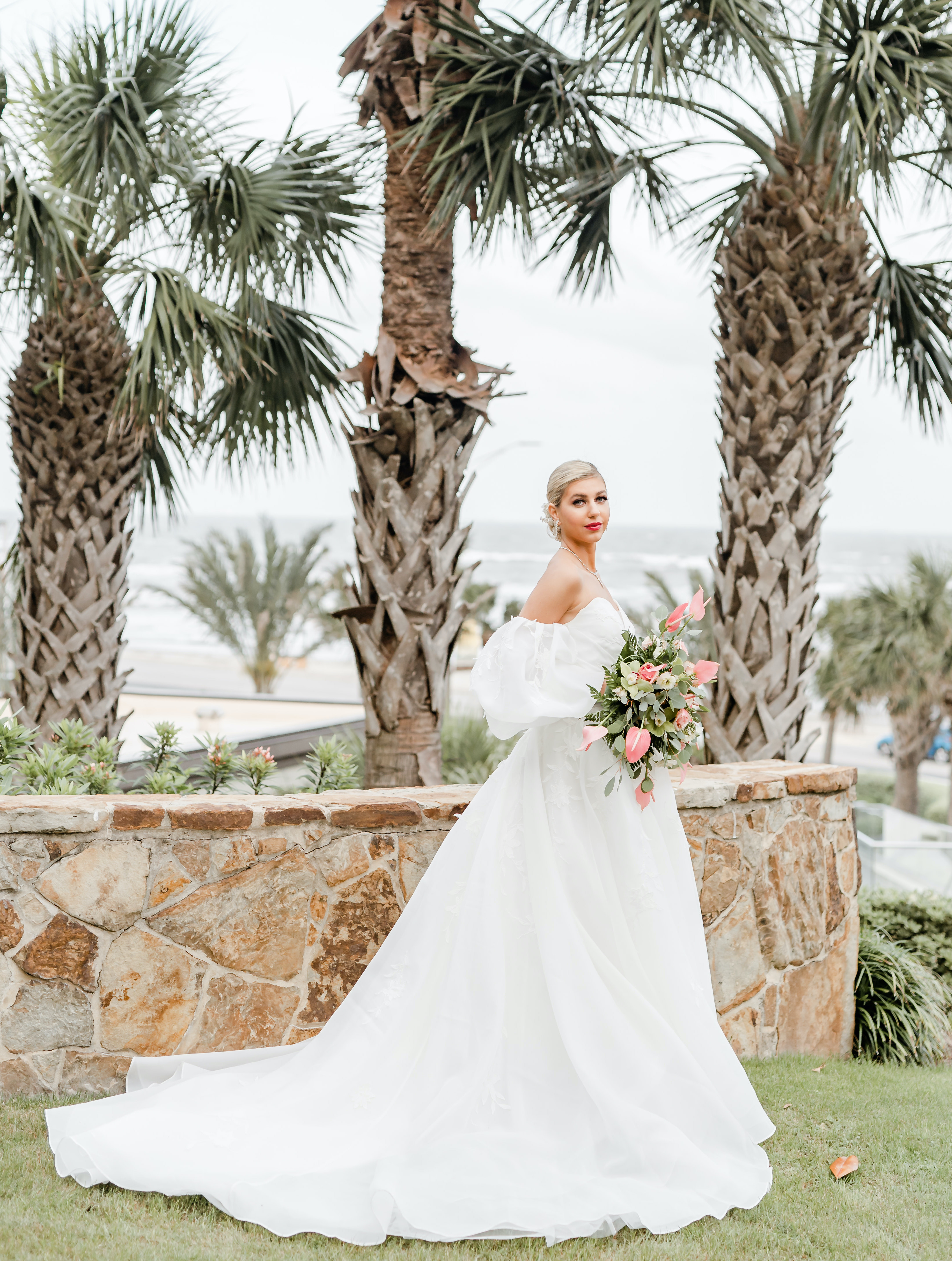 A bride poses in front of the beach and palm trees with a puffy wedding gown and a tropical bridal bouquet.