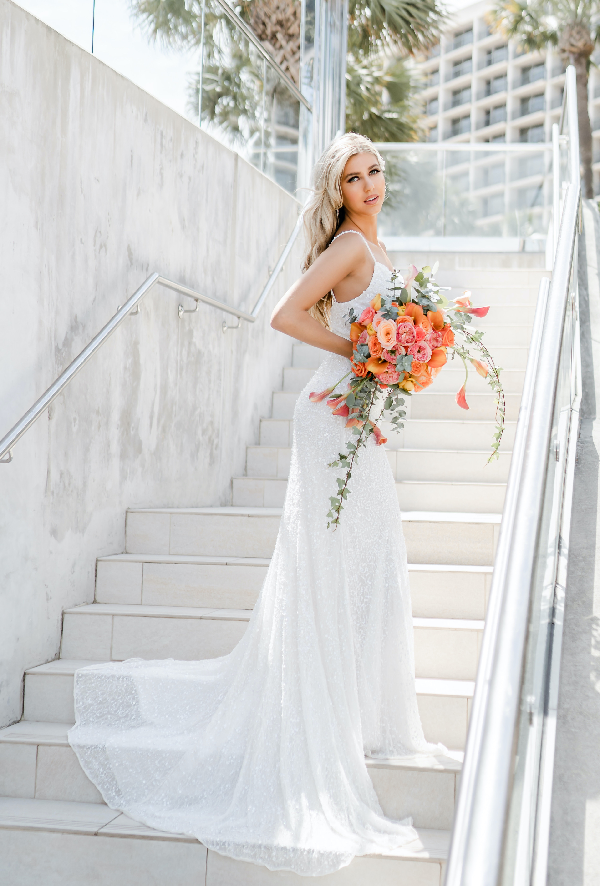 A bride poses on an outdoor stairwell with a bridal bouquet of vibrant orange and pink flowers for a tropical luxe beachfront wedding editorial at The San Luis Resort, Spa & Conference Center in Galveston.