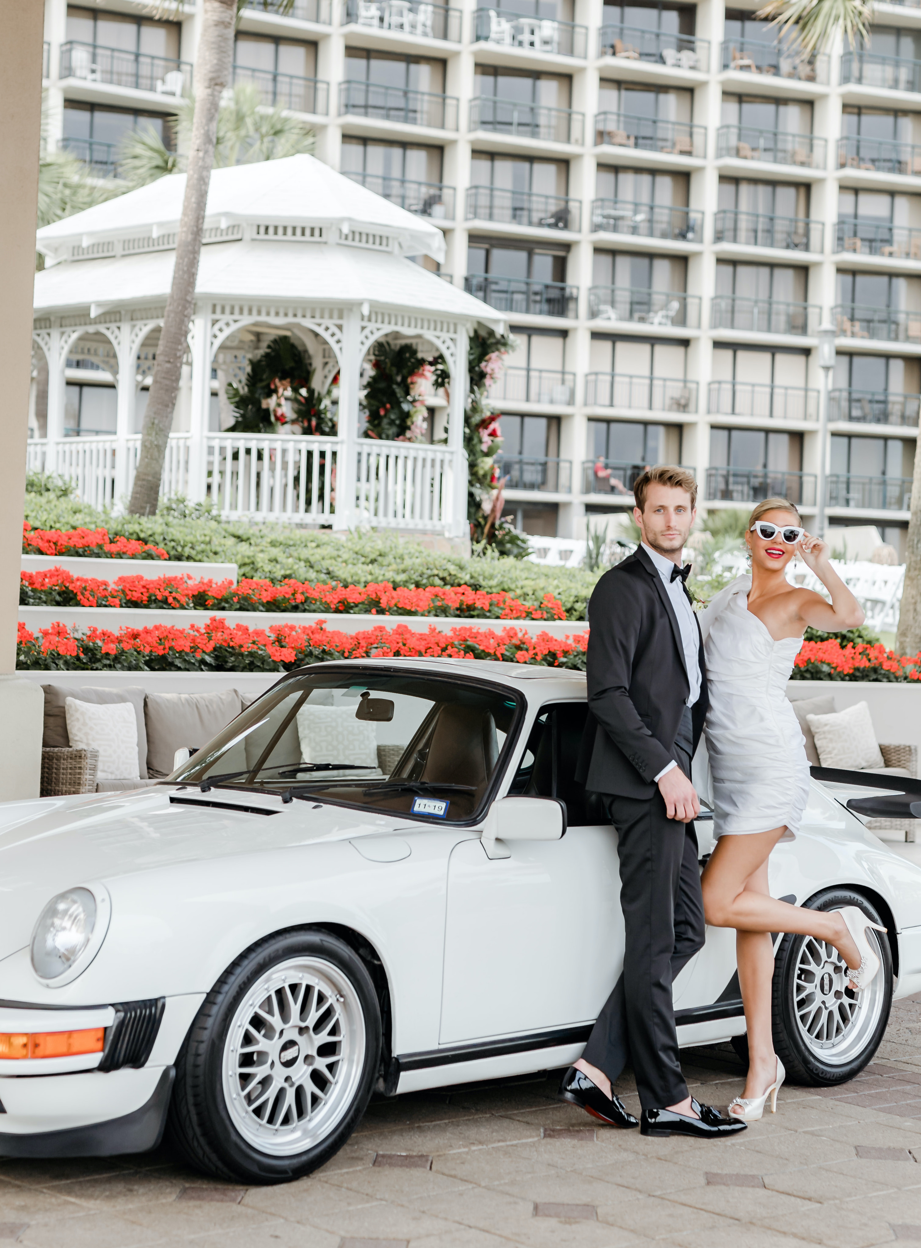 A bride and groom stand in front of a white Porsche that is parked outside of The San Luis Resort, Spa & Conference Center.