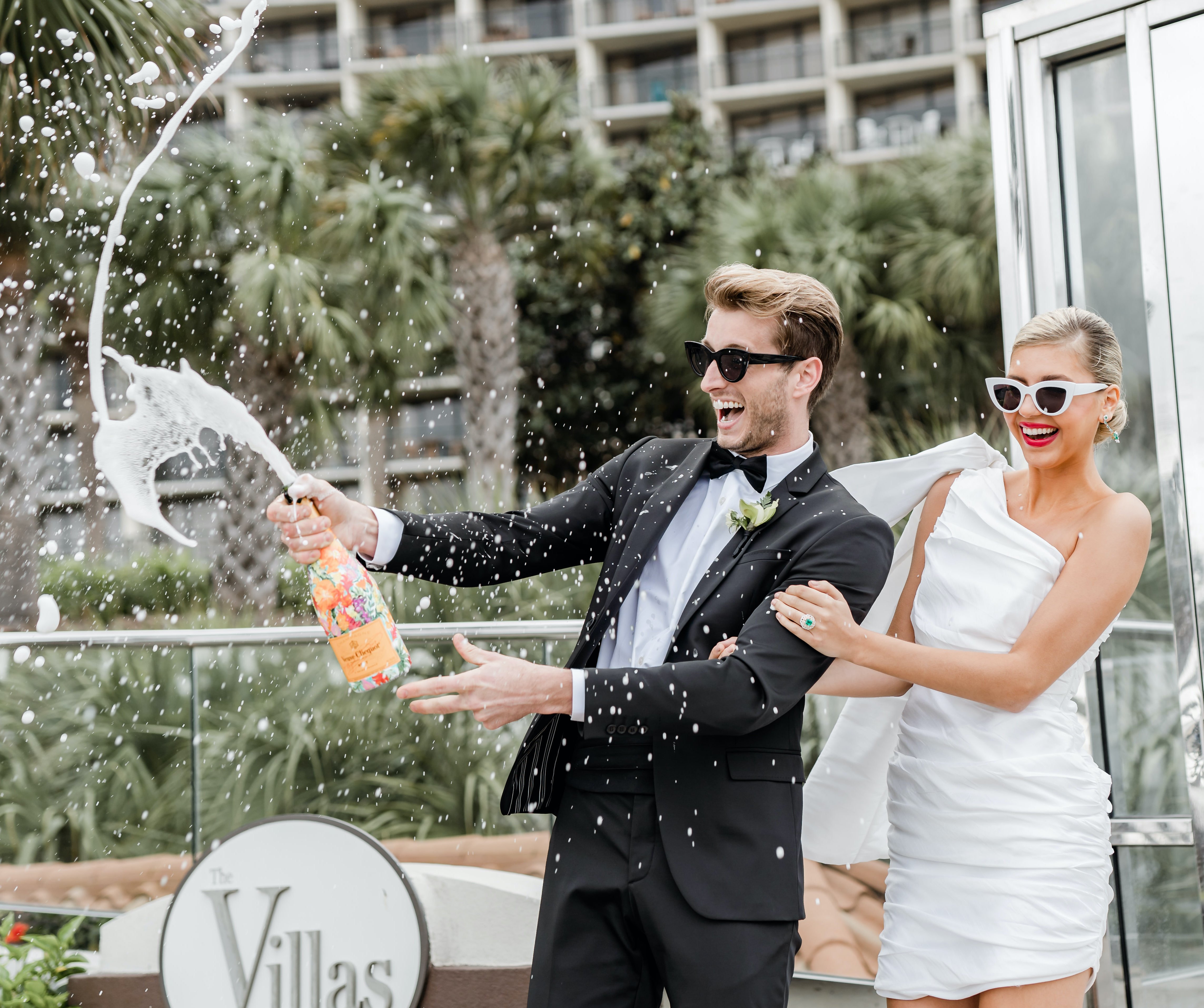 A groom sprays champagne in the air while the bride smiles and watches. They are standing outside at The San Luis Resort, Spa & Conference Center in Galveston, TX.