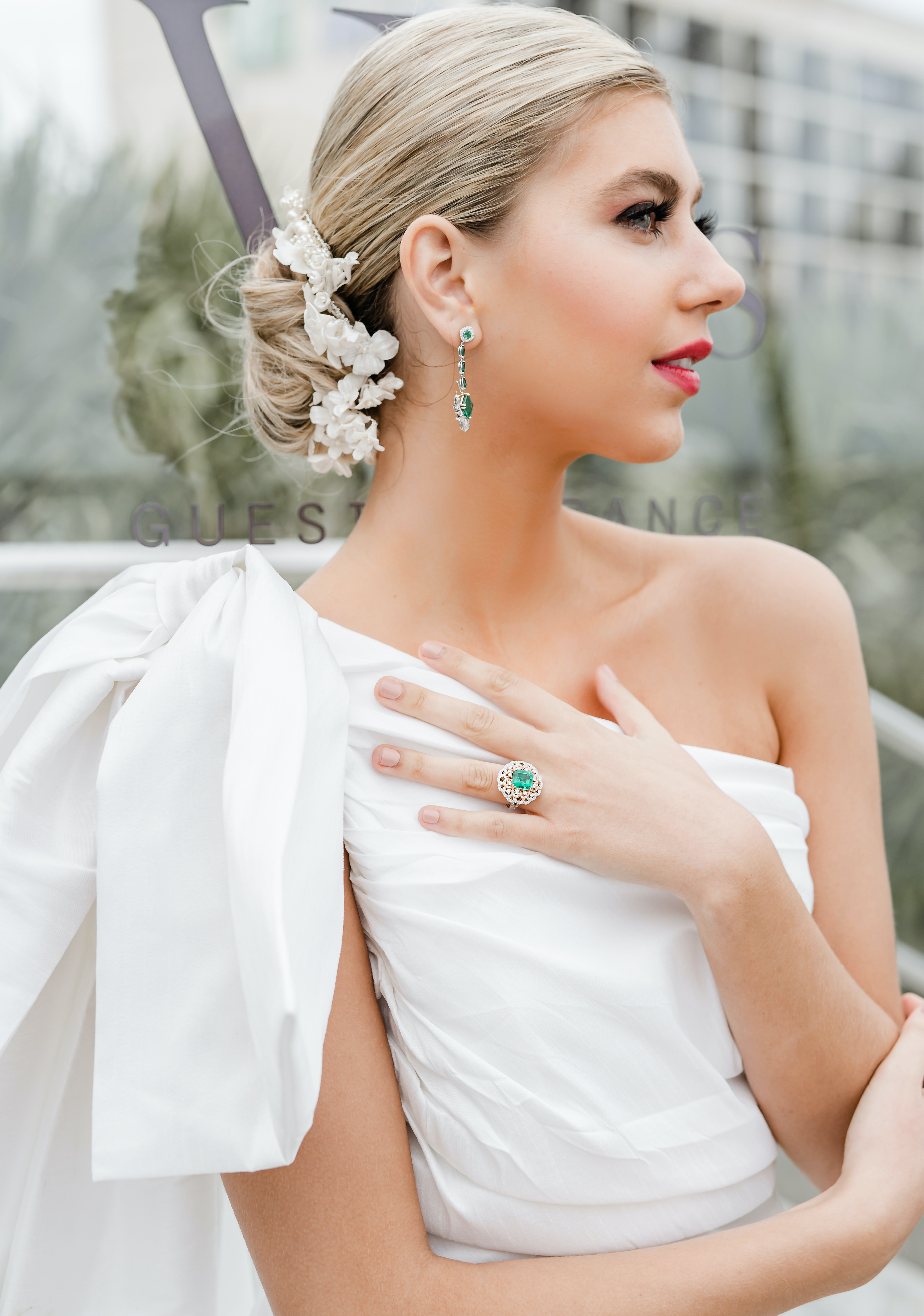 A bride looks away while showing her emerald engagement ring to the camera. Her hair is in a bun with a white flower comb sticking out the side.