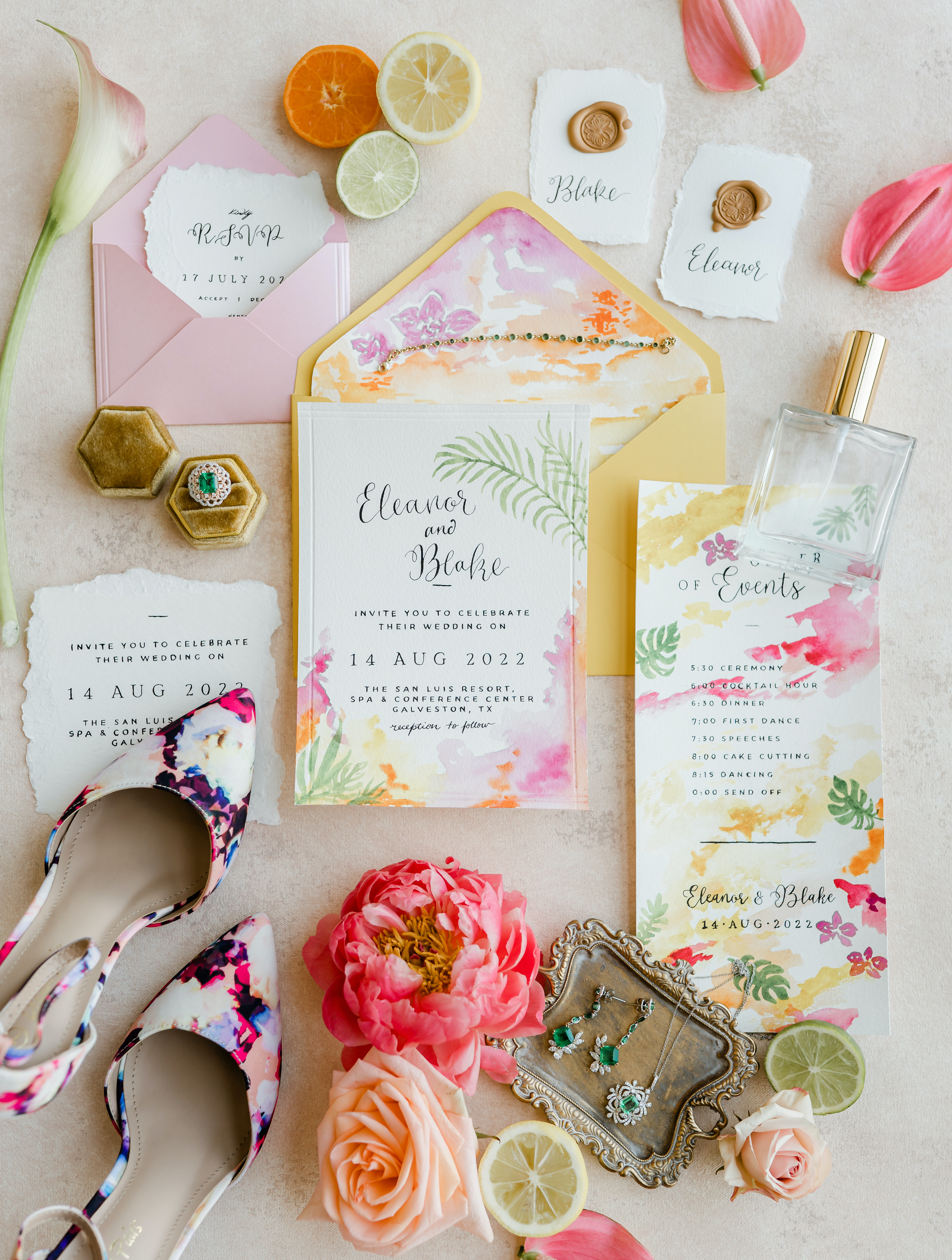 A flatlay of a tropical watercolor invitation suite with bright orange and pink flowers with citrus surrounding it.