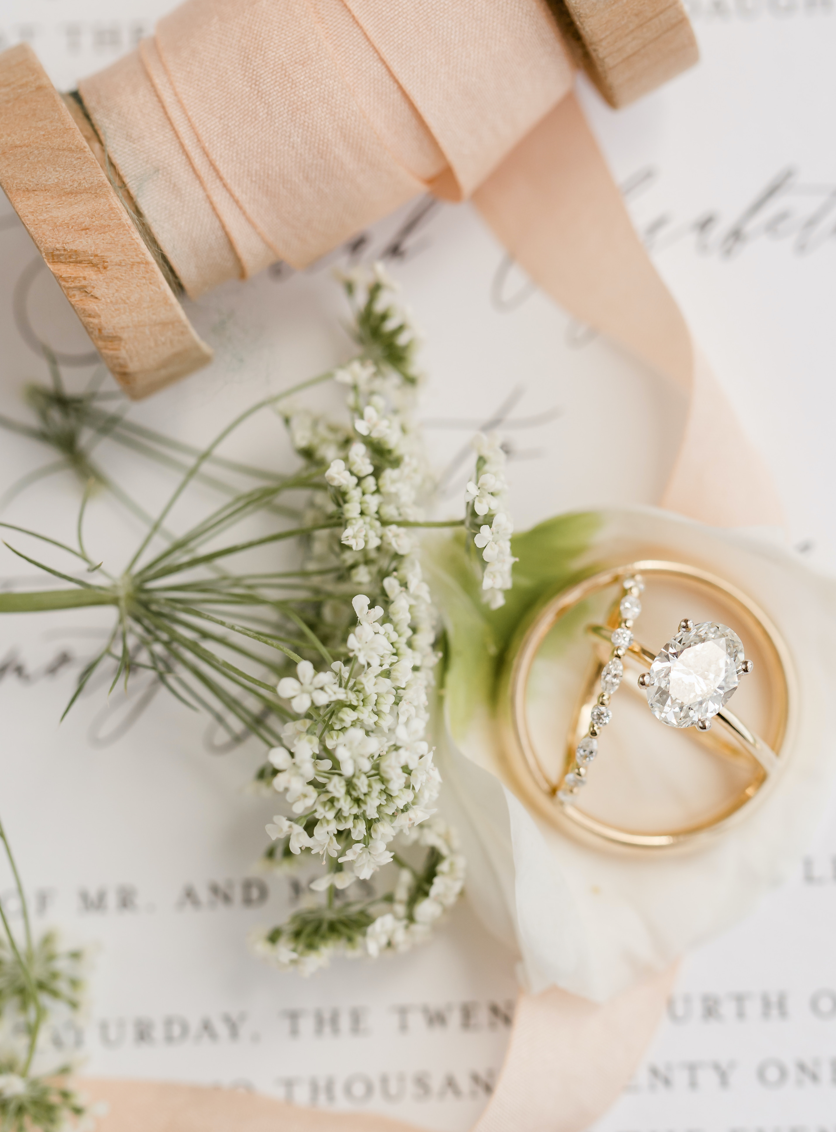 A flat lay of an oval engagement ring surrounded by a spool of blush ribbon and a piece of Queen Anne's Lace.