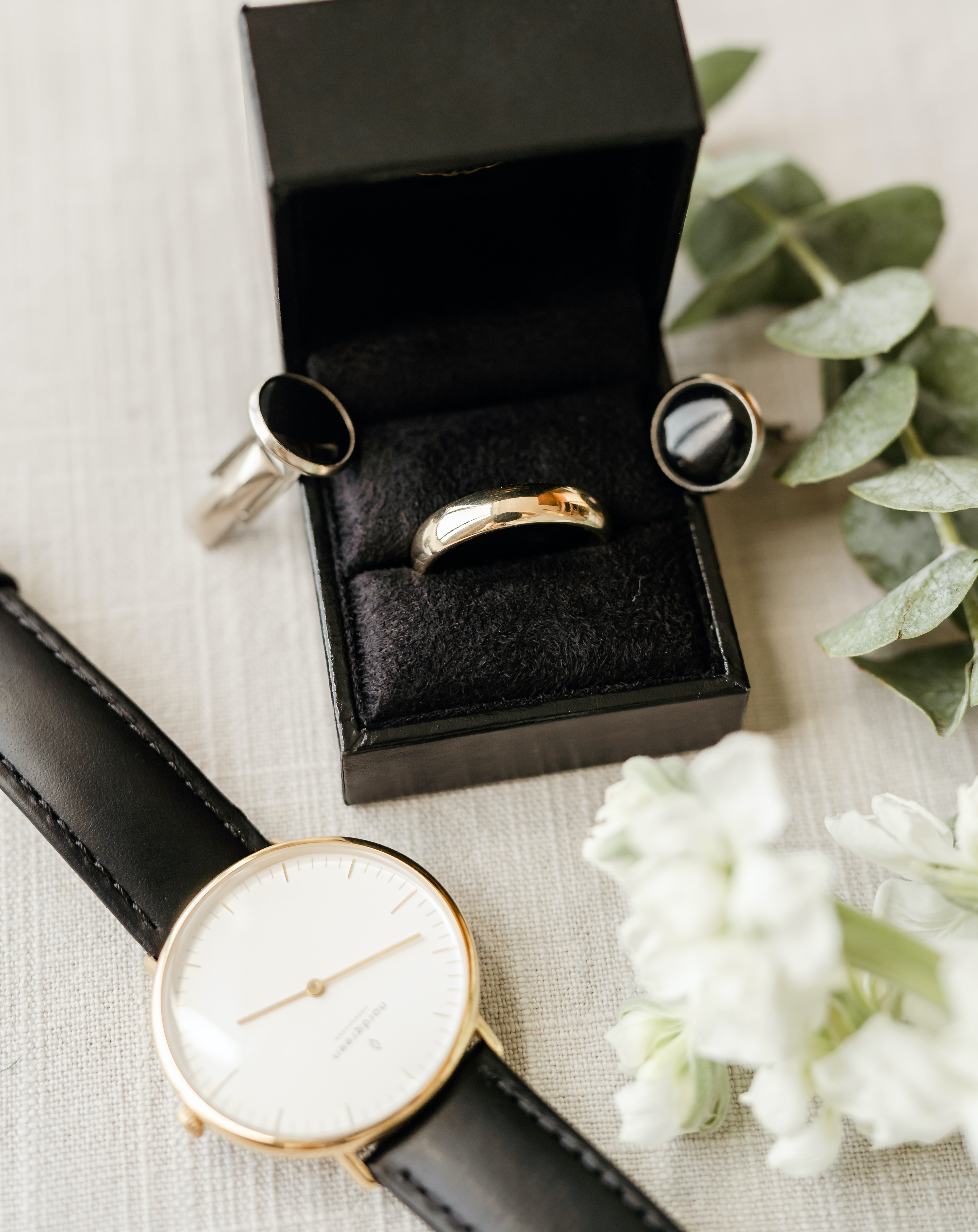 A groom's wedding band sits in a black box surrounded by his walk and a piece of Eucalyptus and white stock.