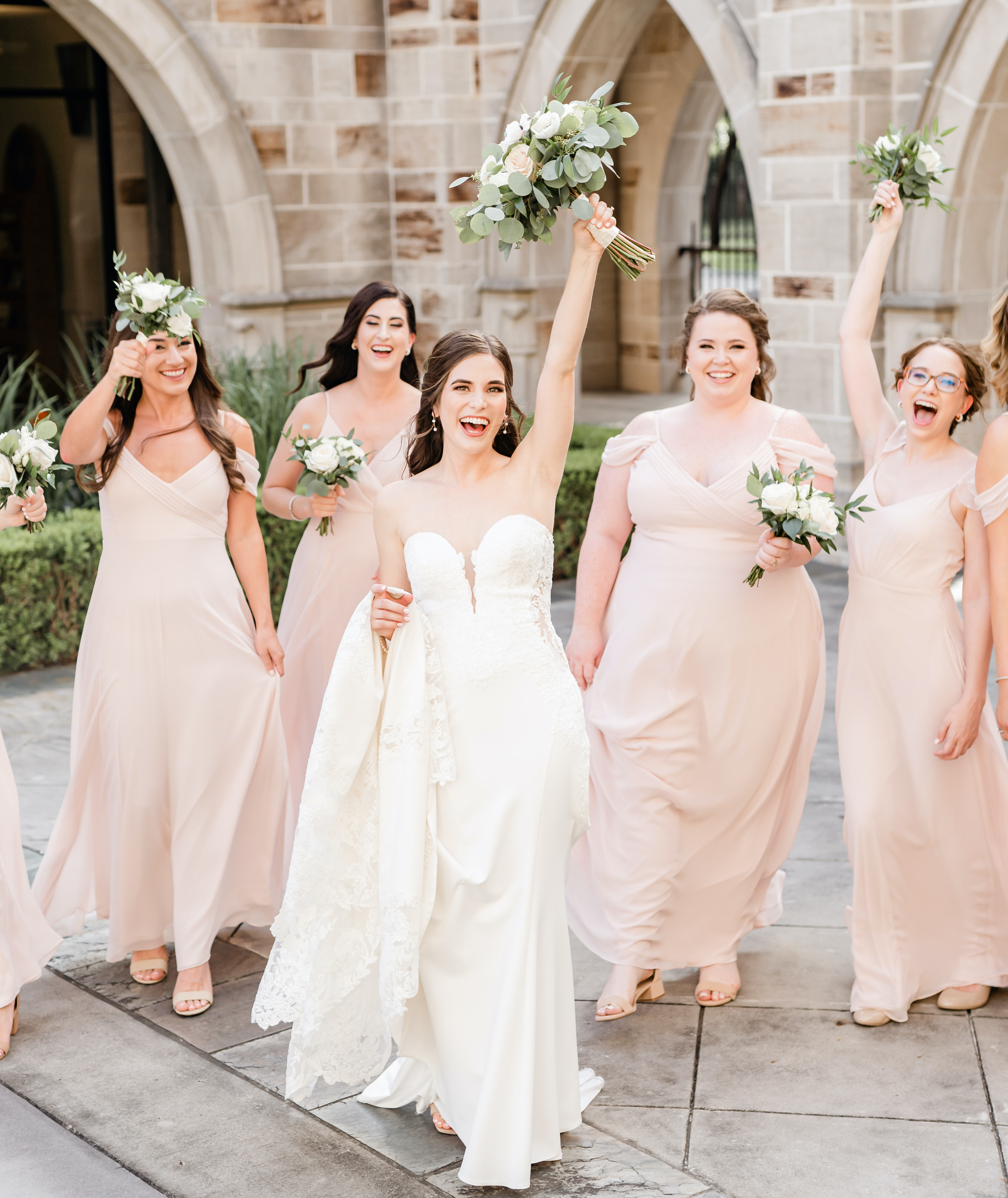 A bride holds her bouquet in the air while her bridesmaids follow behind her.