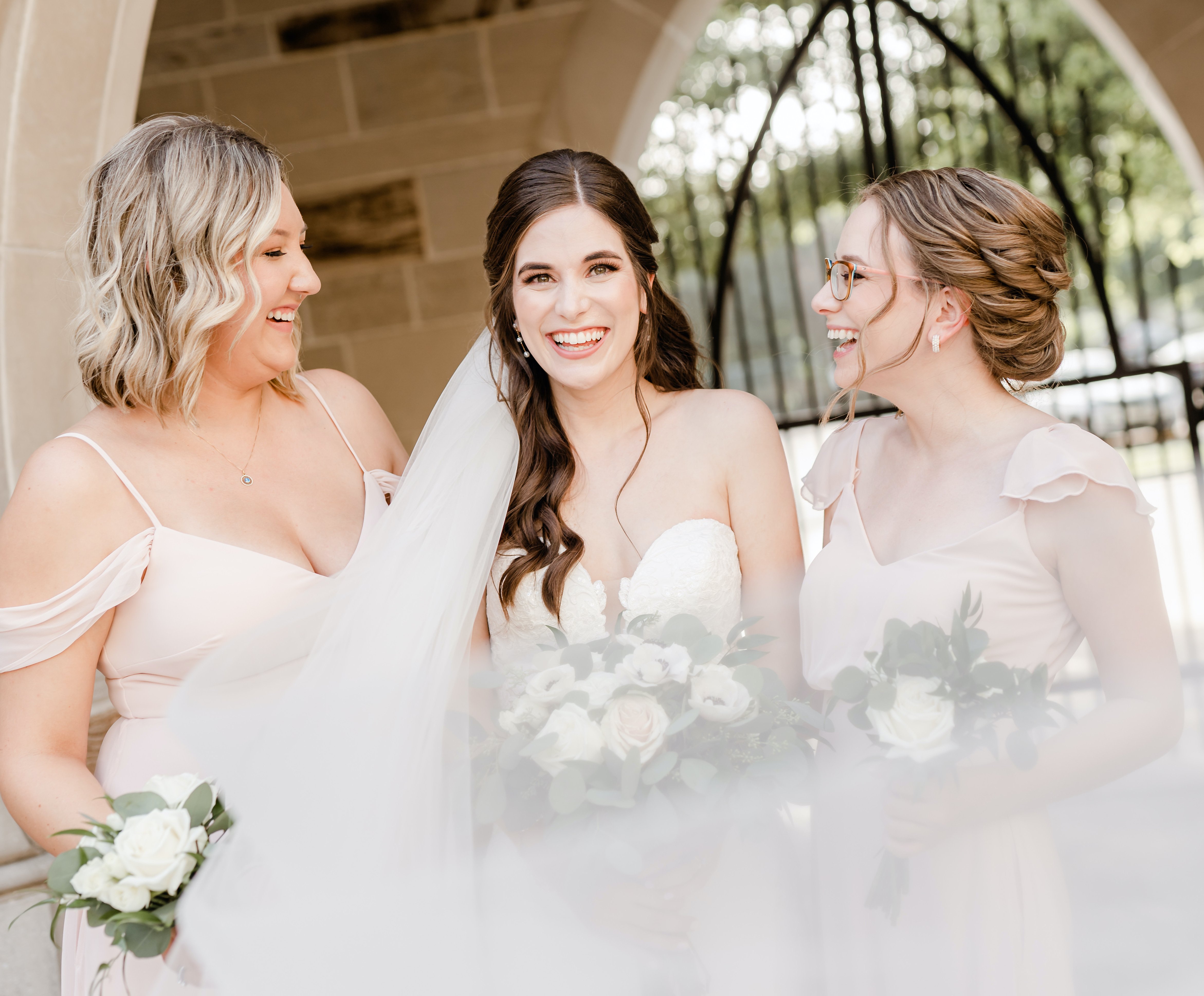A bride stands in the middle of two of her bridesmaids while smiling and laughing outside.
