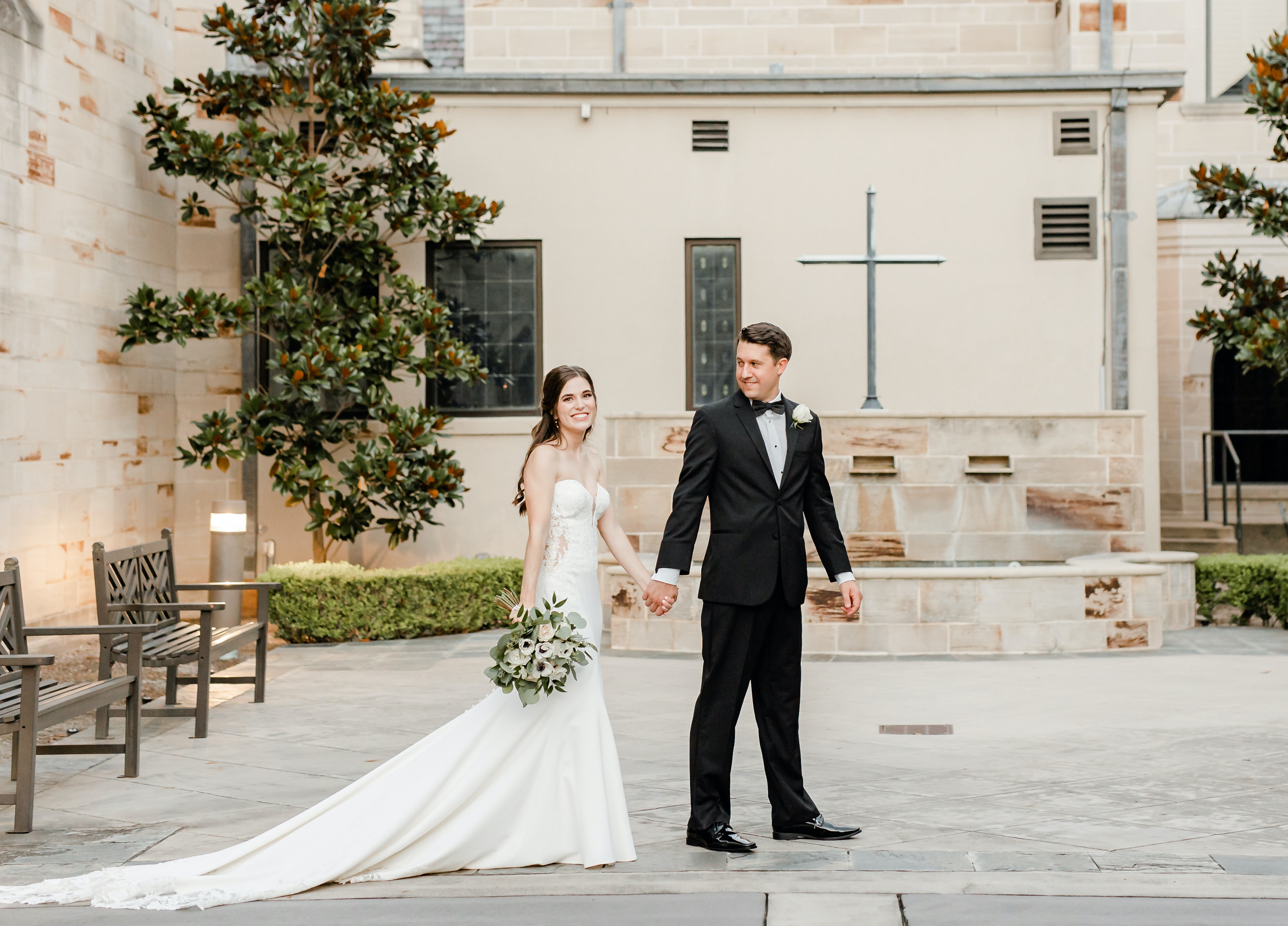 A groom holds hands with his bride as they walk in a courtyard at a church in Houston.