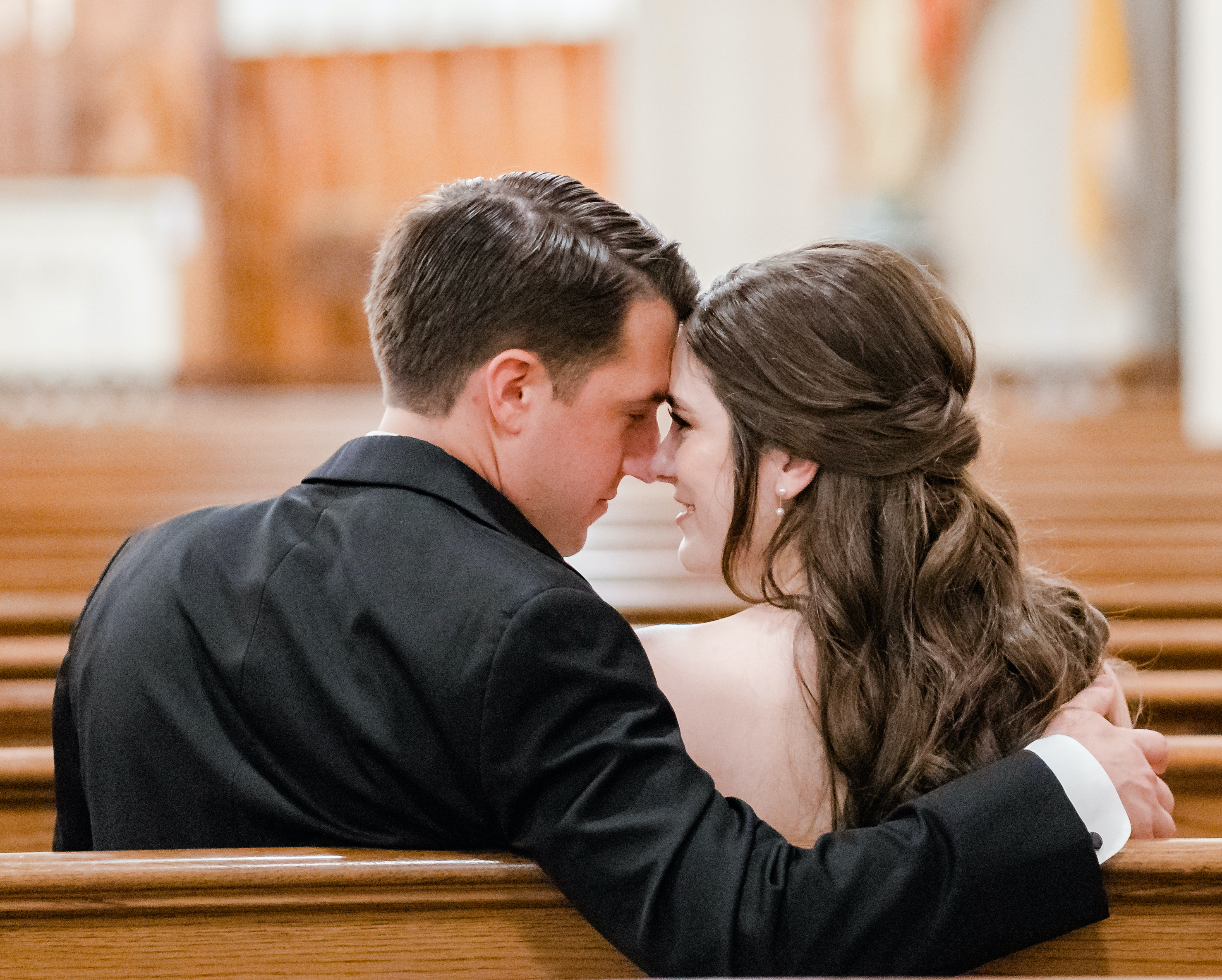 A groom puts his arm around his bride and they touch noses while sitting in a pew in the church.