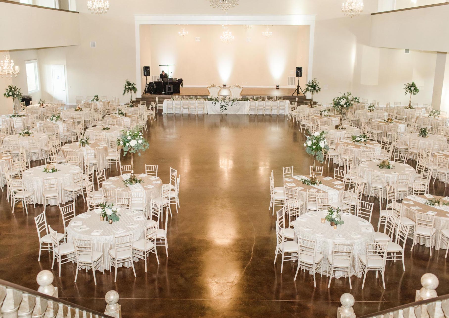 An open reception room with white chairs and table cloths and greenery centerpieces set up for a wedding at The Hundred Oaks Event Center in Houston, TX.