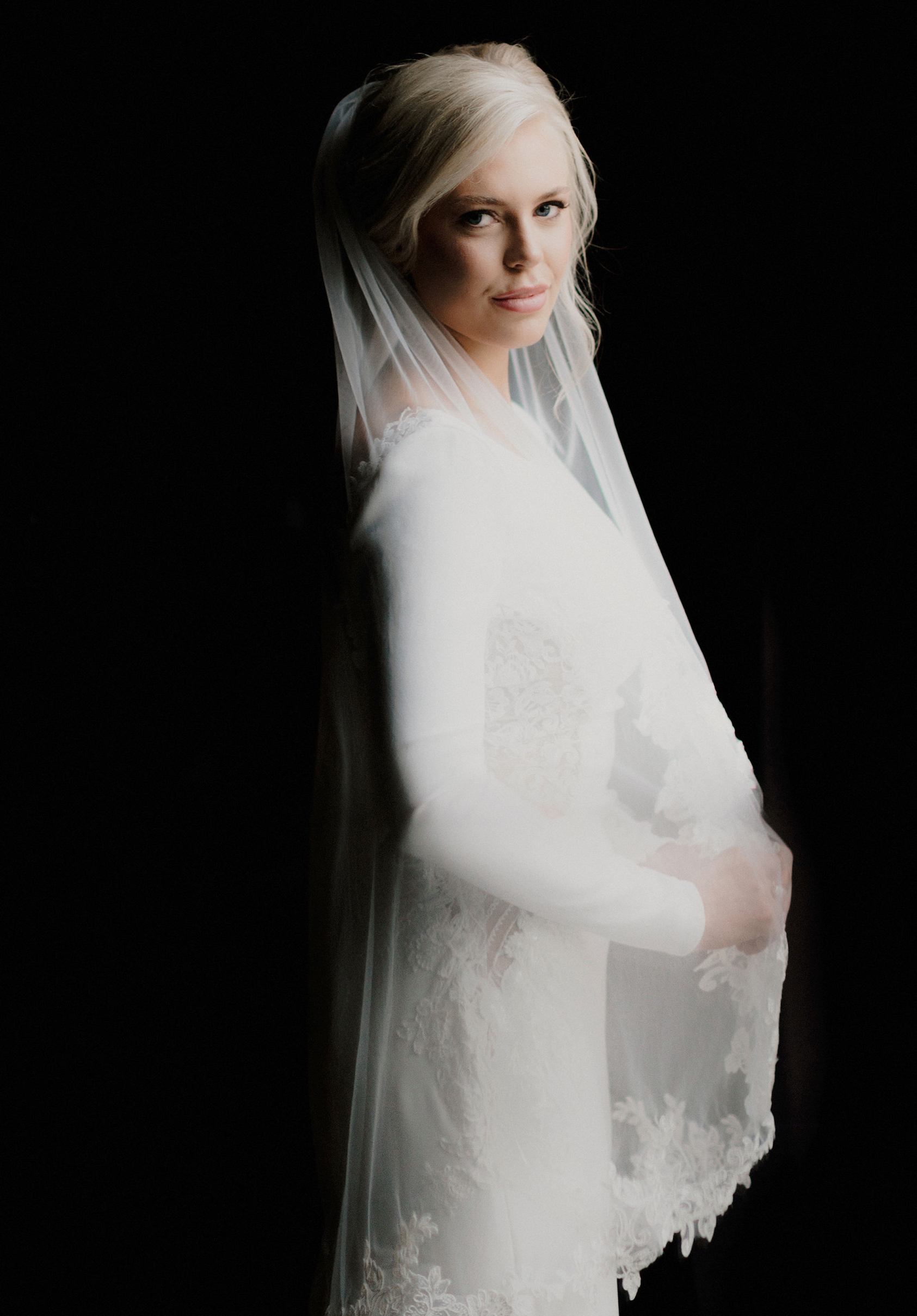 A bride wears her wedding gown and veil against a black background.