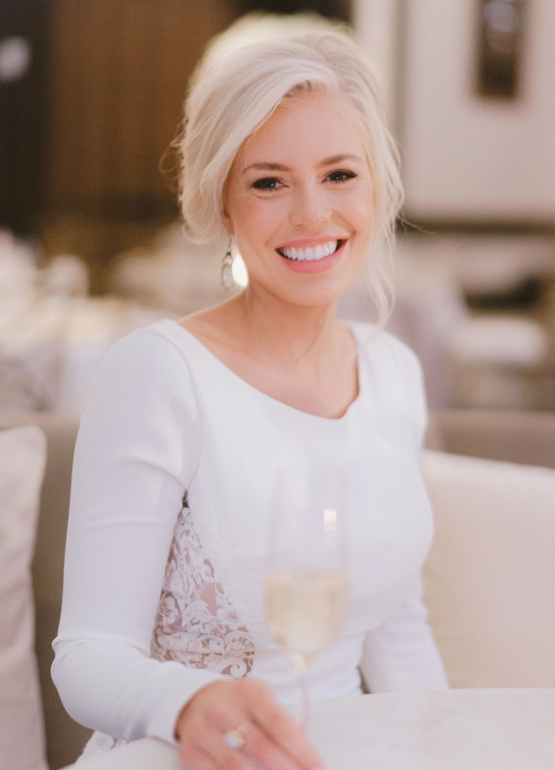 A bride smiles with her hair pulled back and wearing a longsleeve dress with lace cutouts on the sides for her romantic ballroom wedding.