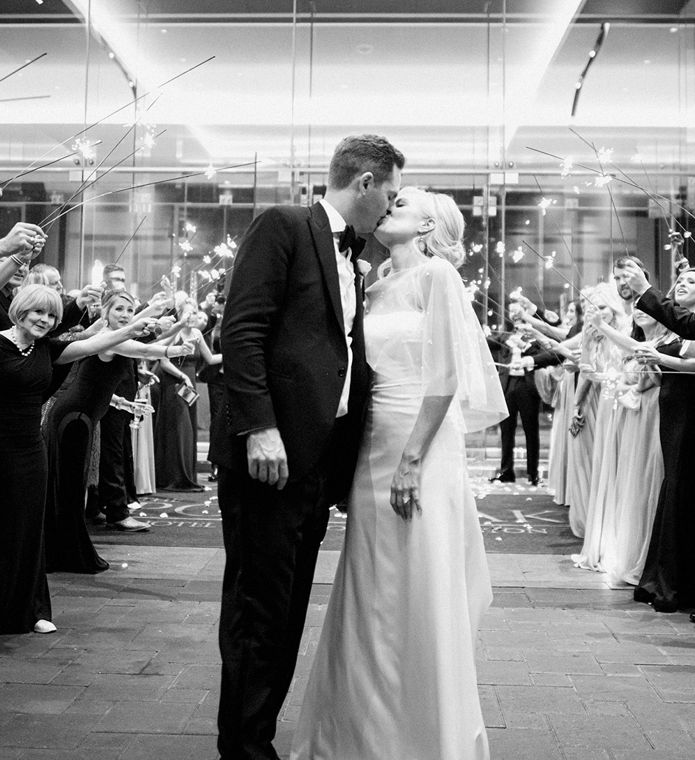 A black and white photo of a bride and groom kissing in the middle of the send-off aisle their freinds made.