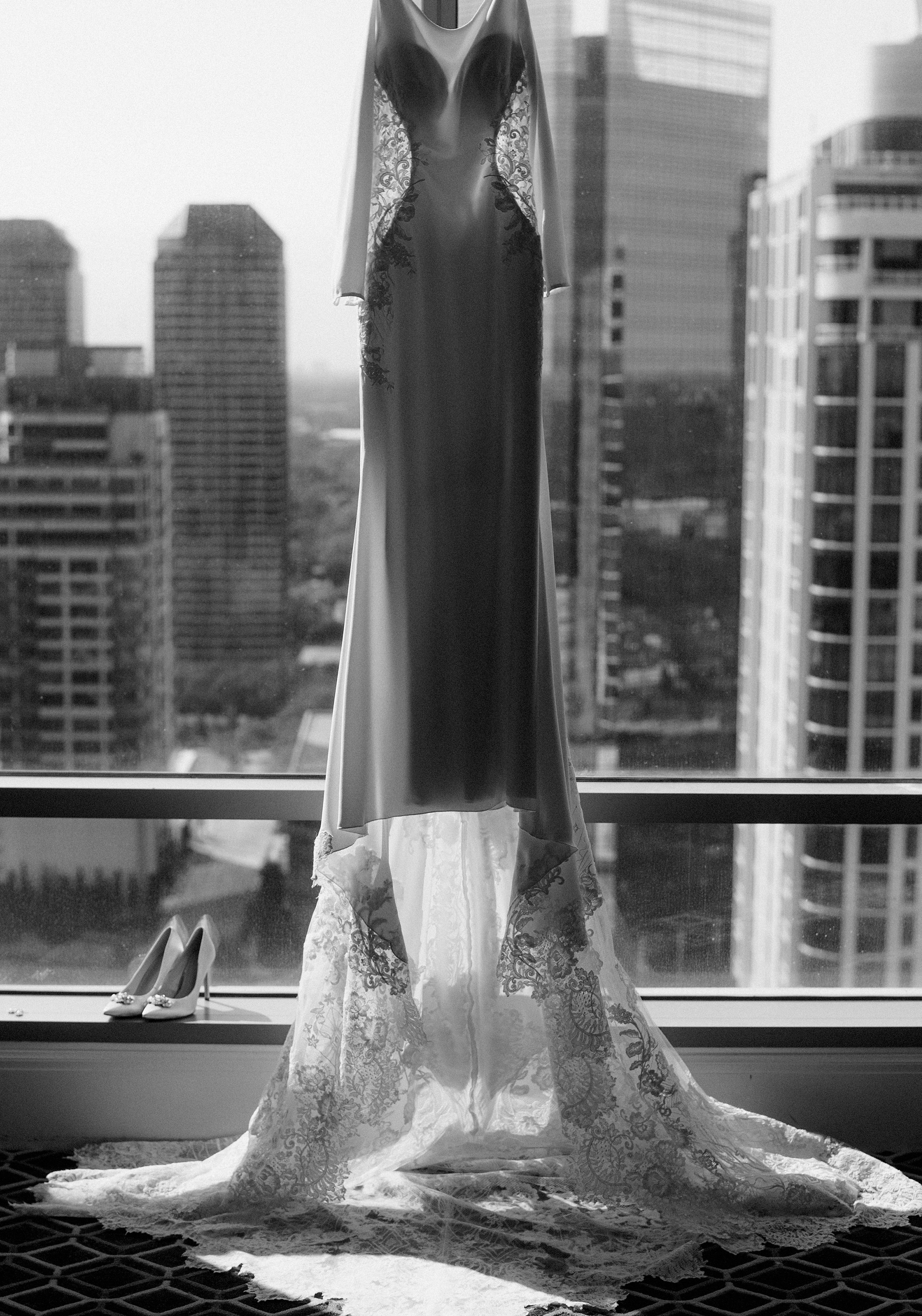 A black and white photo of a bride's wedding dress hanging from a hotel window overlooking the Houston skyline.